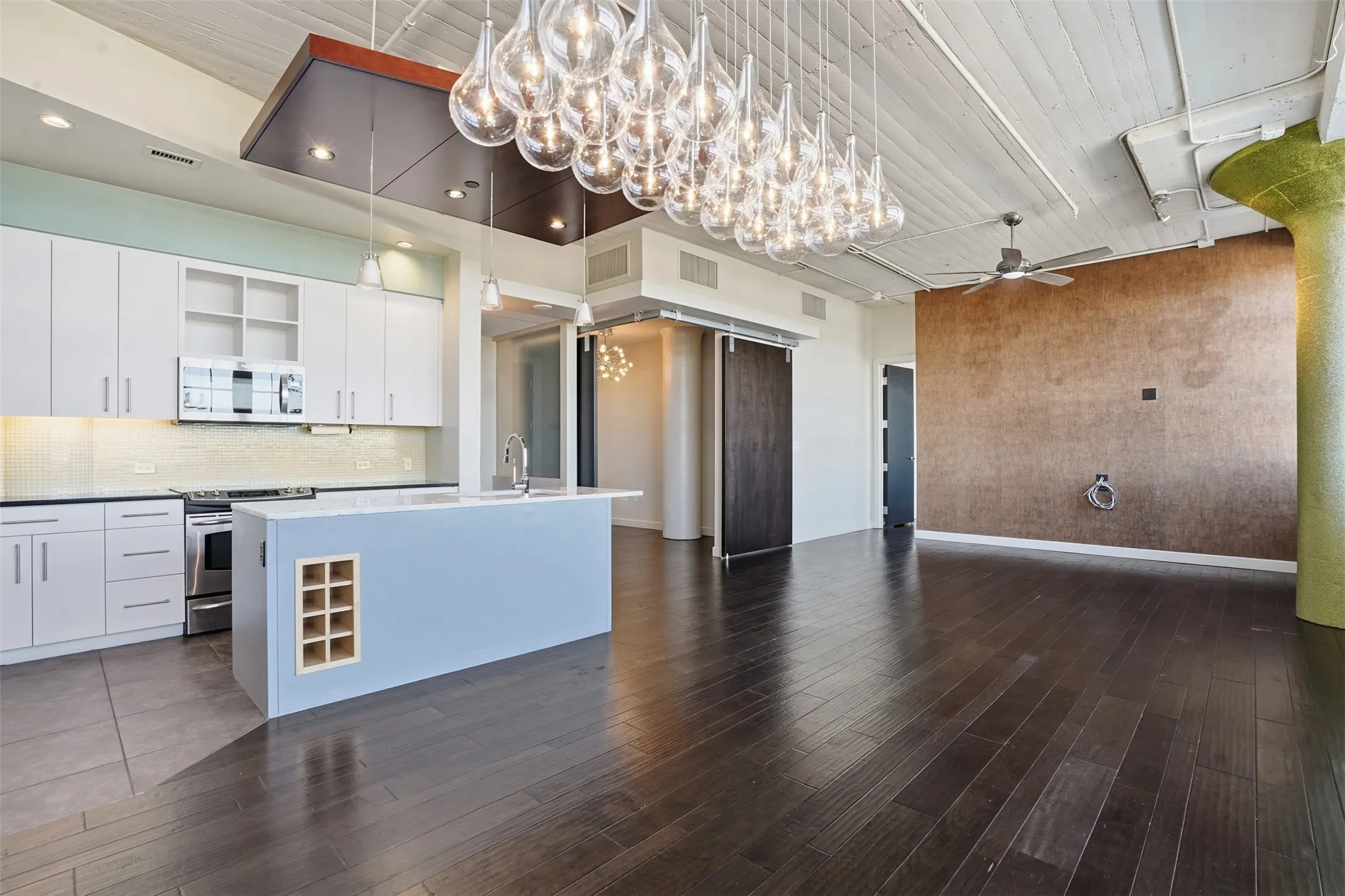Open-concept living area featuring dark wood-finish flooring, a contemporary kitchen island with a built-in wine rack, a stainless steel range and microwave, and extensive overhead lighting fixtures