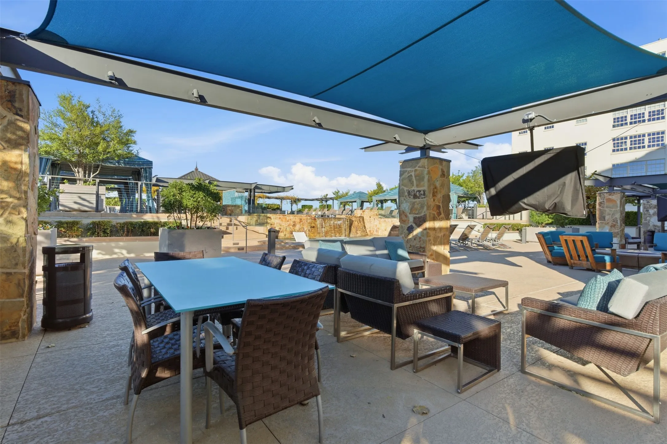 Expansive outdoor patio featuring a large blue fabric shade structure, stone-clad support columns, and durable concrete paving