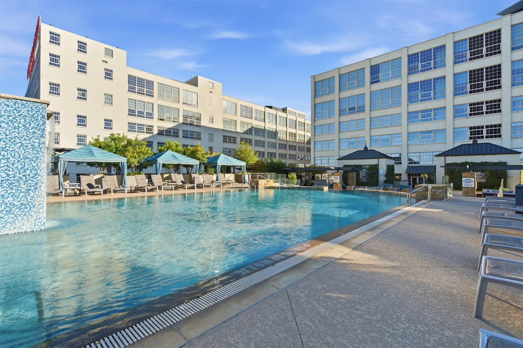 Expansive outdoor swimming pool featuring a tiled waterfall element, multiple shaded cabanas, and sun loungers