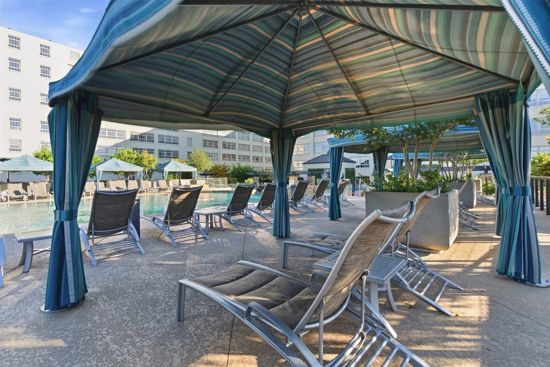 Poolside cabana featuring a striped canopy, privacy drapes, textured concrete patio, and nearby lounge seating