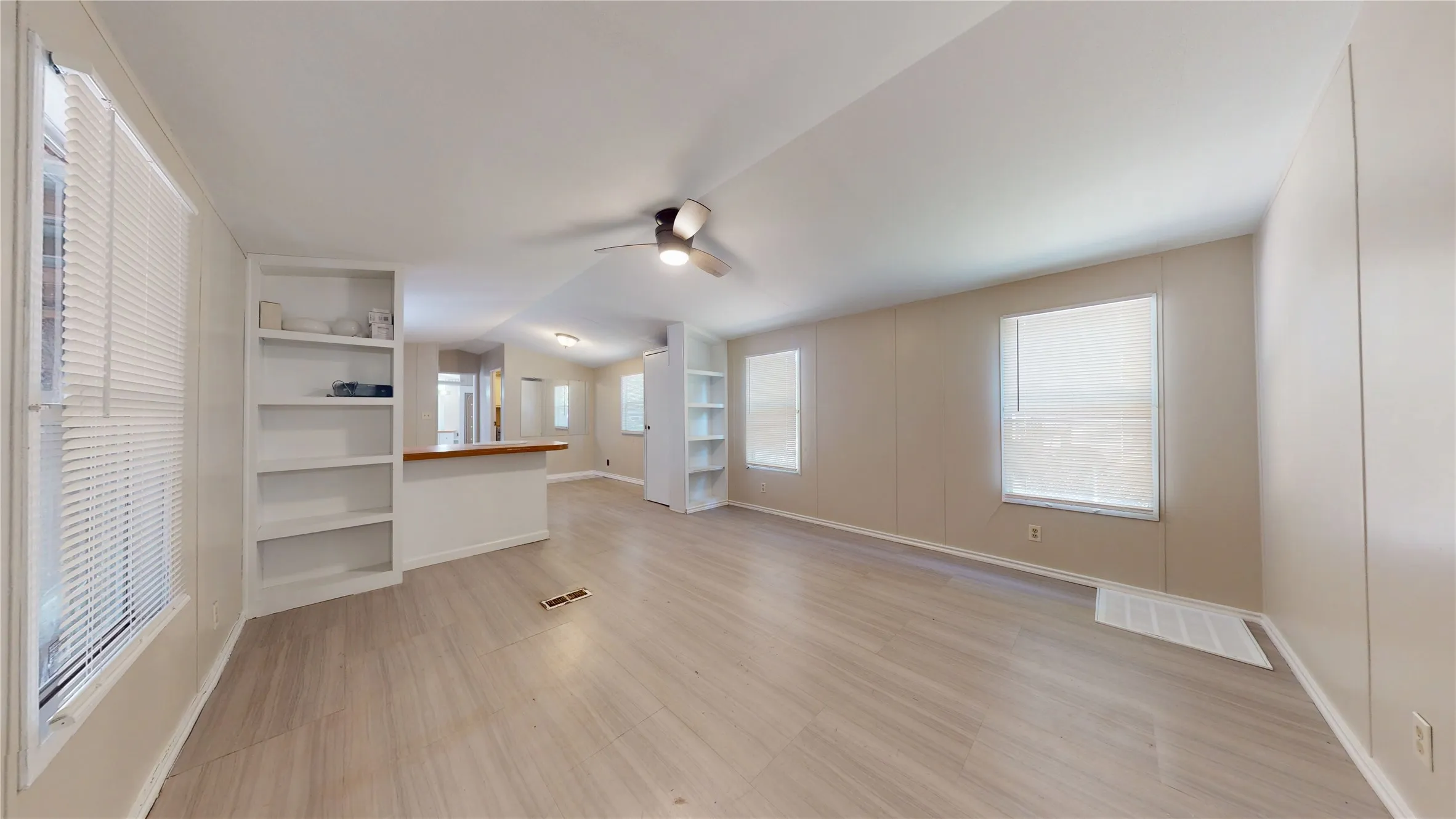 Open-concept living area with wood-finish flooring, built-in shelving, a ceiling fan, and multiple windows with blinds