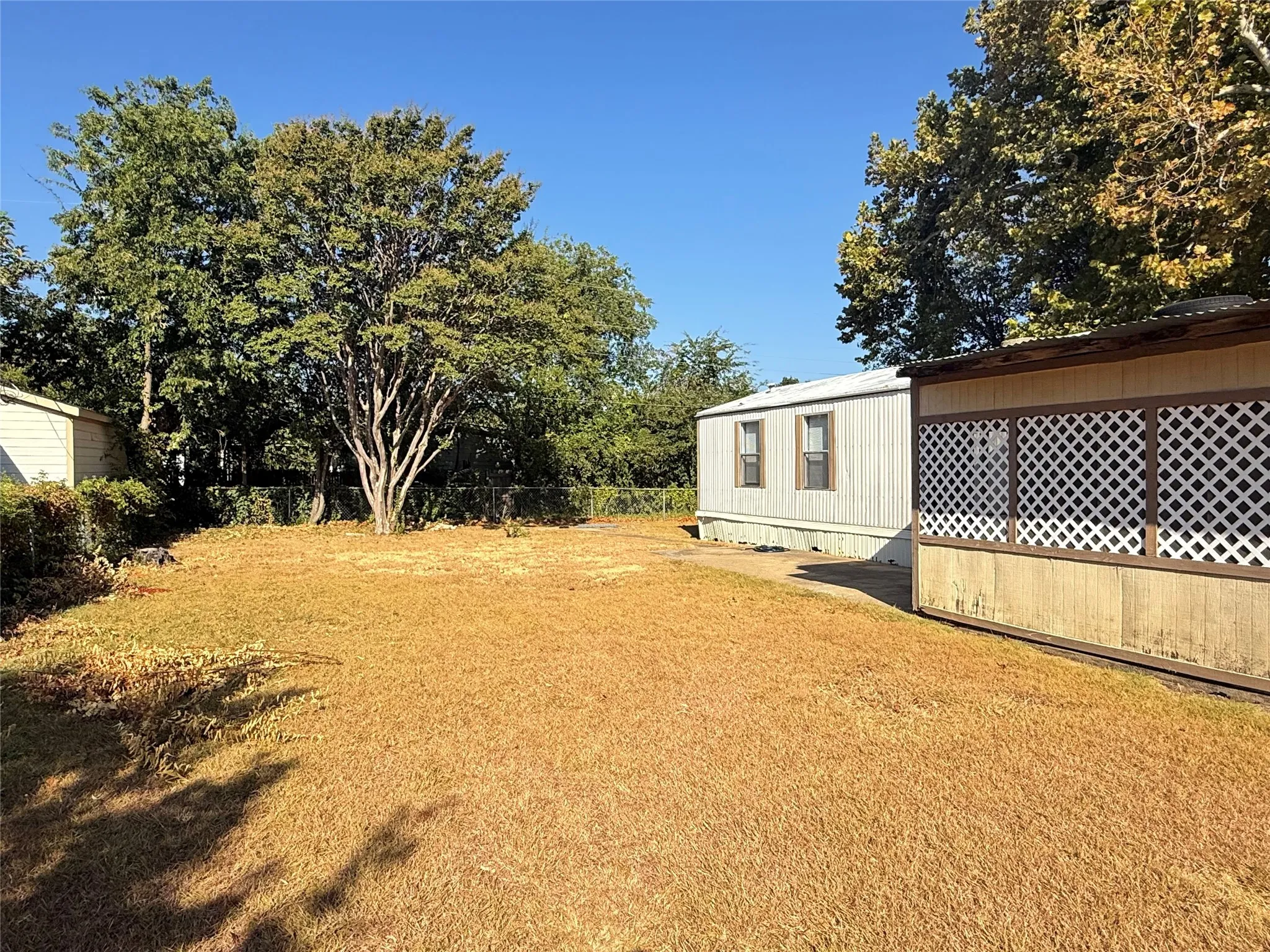 Expansive outdoor space featuring a detached building with lattice detailing, mature trees, and a clear sky backdrop