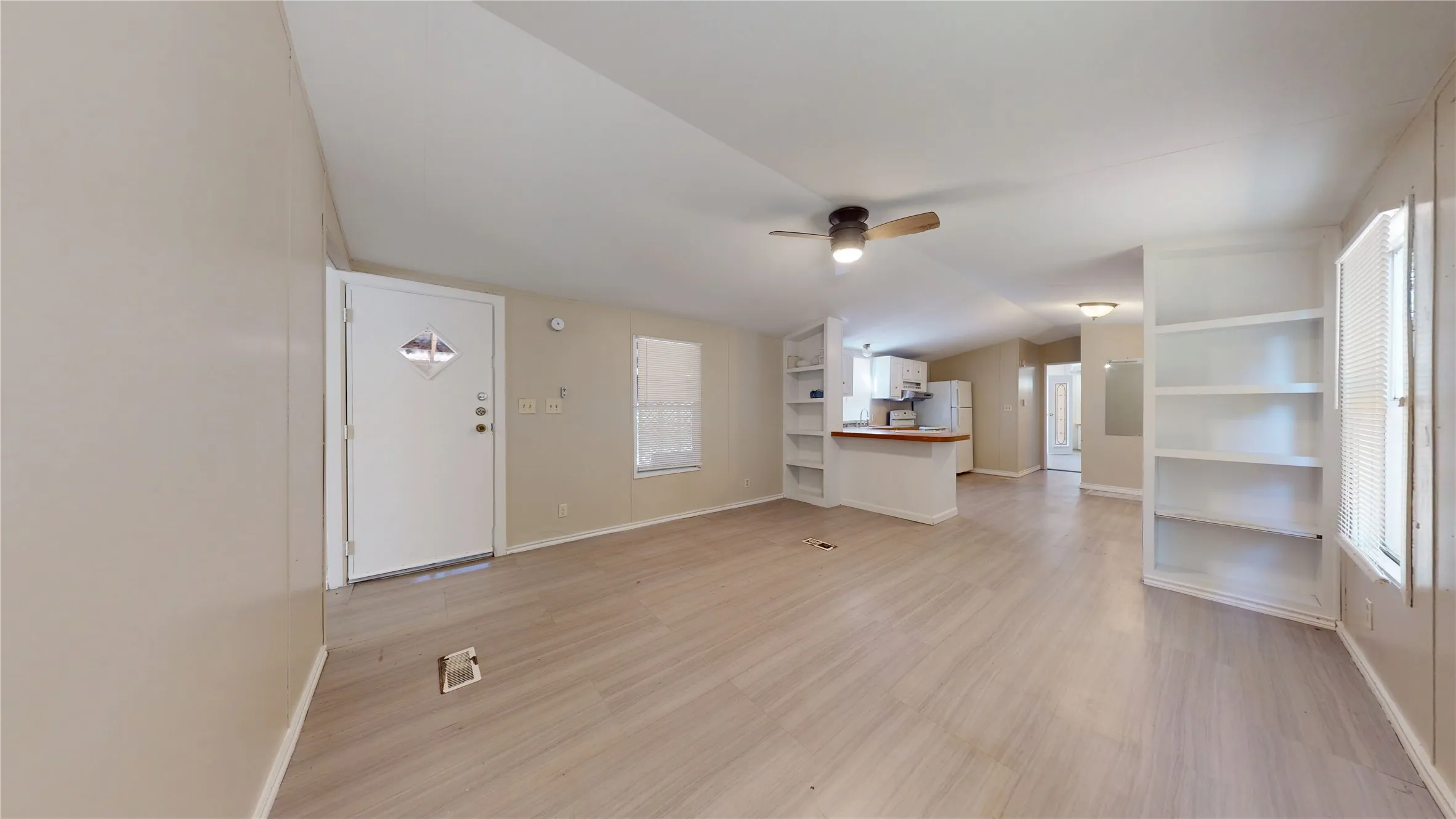 Light wood-finish flooring, built-in shelving, white entry door with decorative glass, ceiling fan with integrated lighting, and a partial kitchen with white appliances