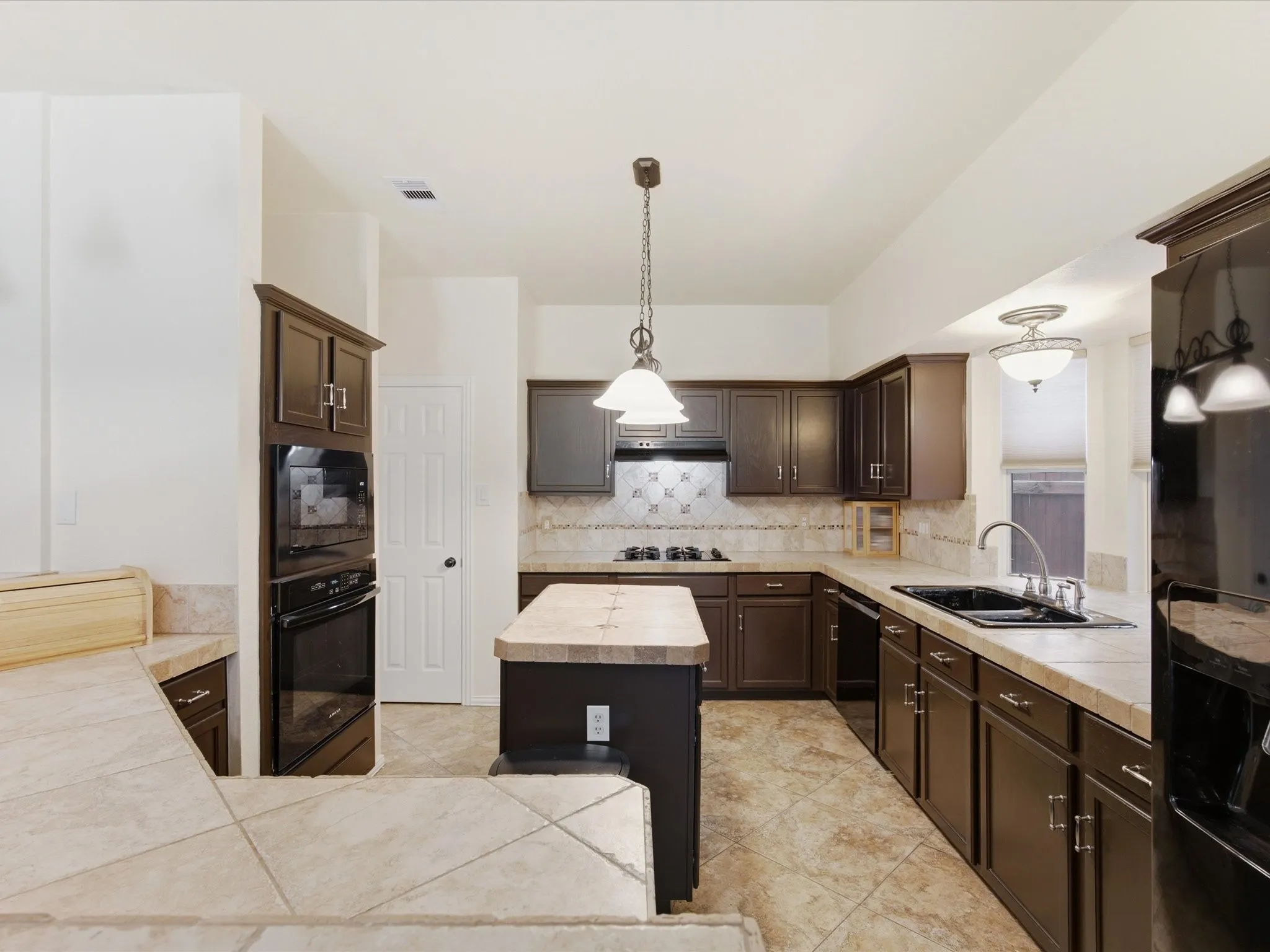Spacious kitchen featuring dark wood cabinetry, tile countertops, a center island with a breakfast bar, stainless steel double oven, and integrated built-in appliances