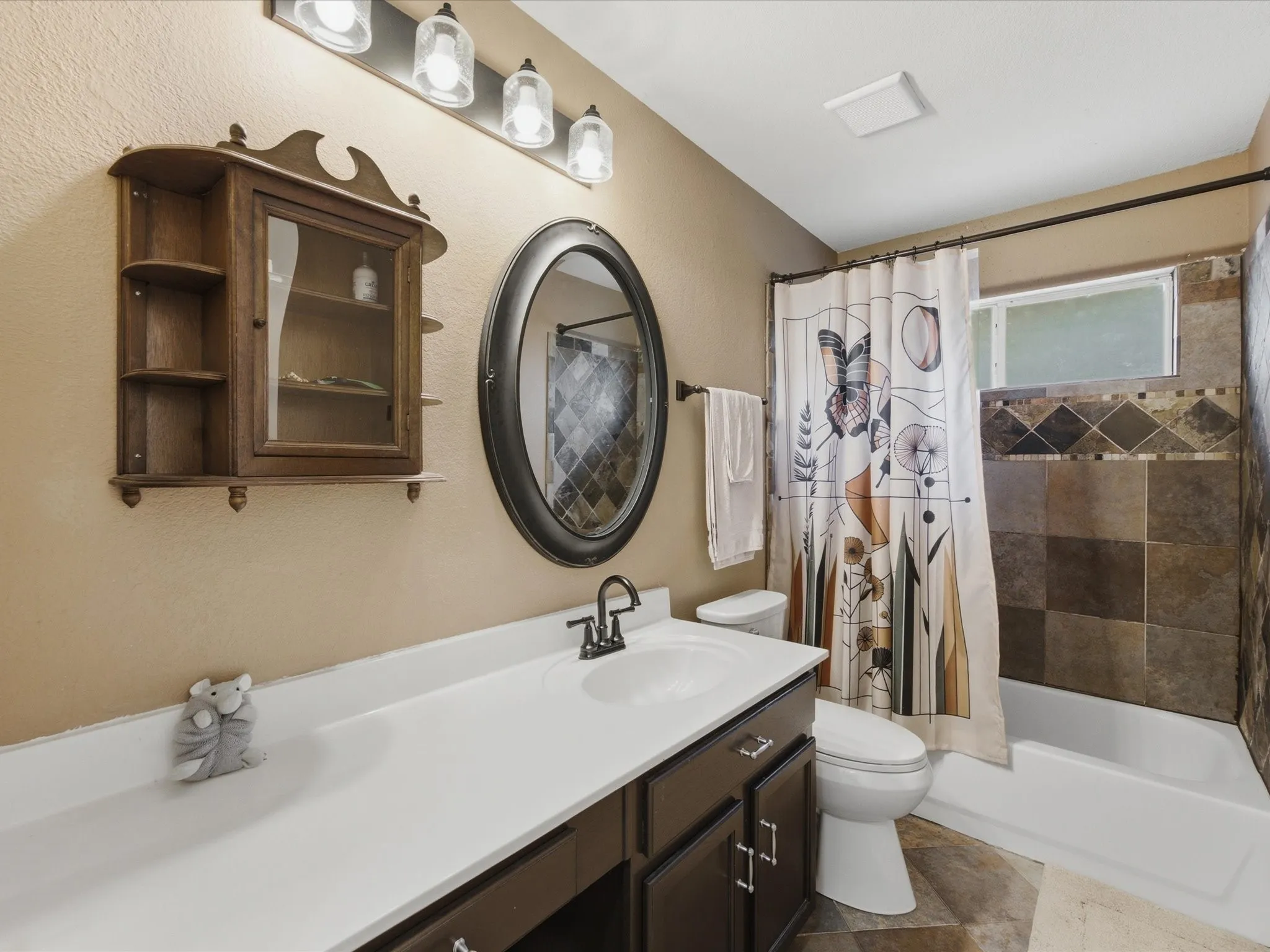 Bathroom featuring an oval mirror, a four-light vanity fixture, a dark wood-finish vanity with a white countertop and integrated sink, a built-in bathtub with a tiled surround, and multi-toned floor tiling