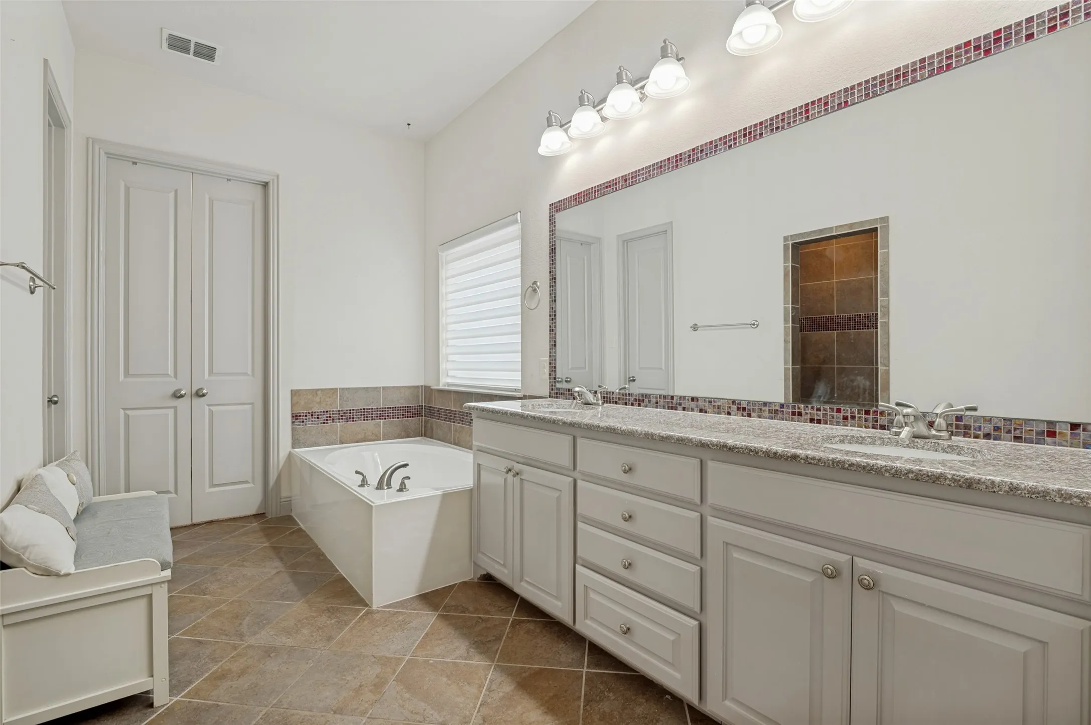 Dual vanity with stone-finish countertop and undermount sinks, featuring a full-width mirror with accent tile border and sconce lighting