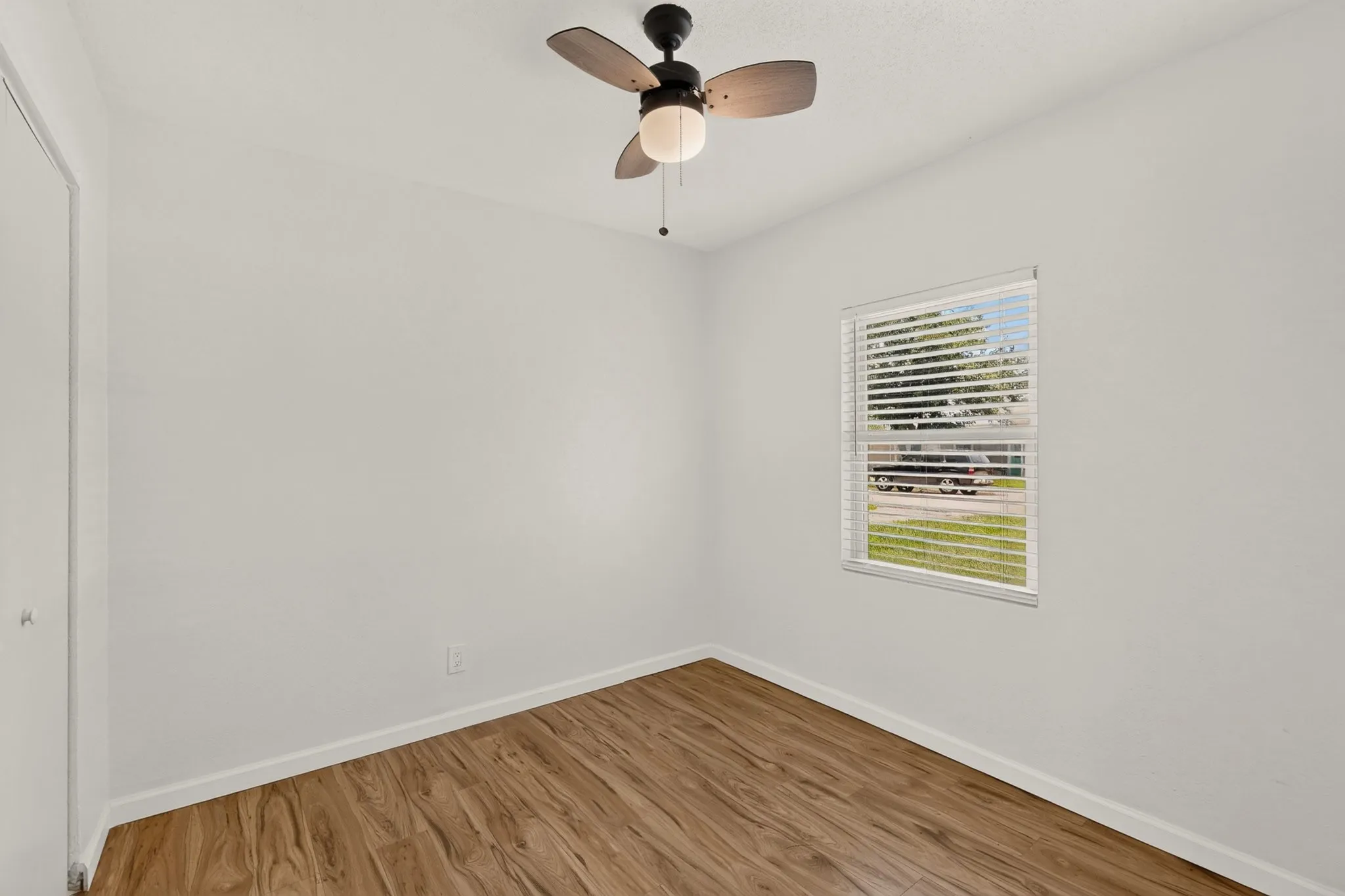 Room featuring light-toned walls, wood-finish flooring, a window with horizontal blinds, a ceiling fan with integrated lighting, and white baseboards