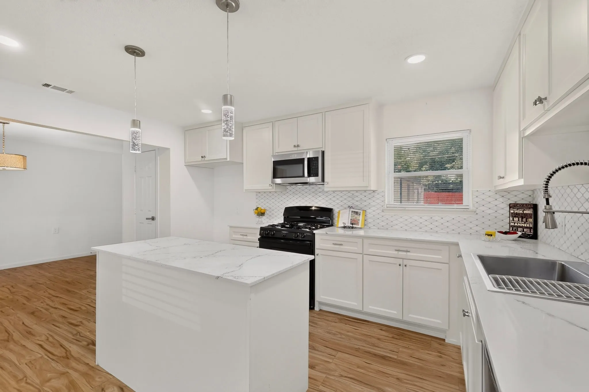 Modern kitchen featuring a central island with a quartz-finish countertop, white cabinetry with silver hardware, a white geometric tile backsplash, a stainless steel sink with a high-arch faucet, and wood-finish flooring
