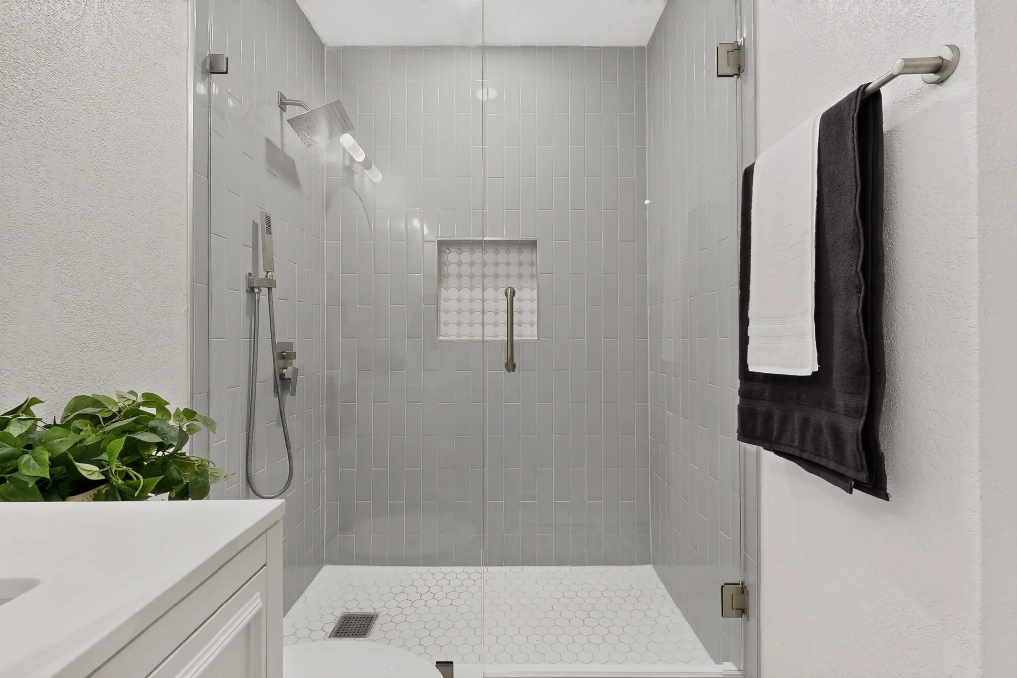 Glass-enclosed shower featuring vertical subway tile walls, a rain shower head, a handheld shower fixture, a recessed niche with decorative tile, and a hexagonal tile floor