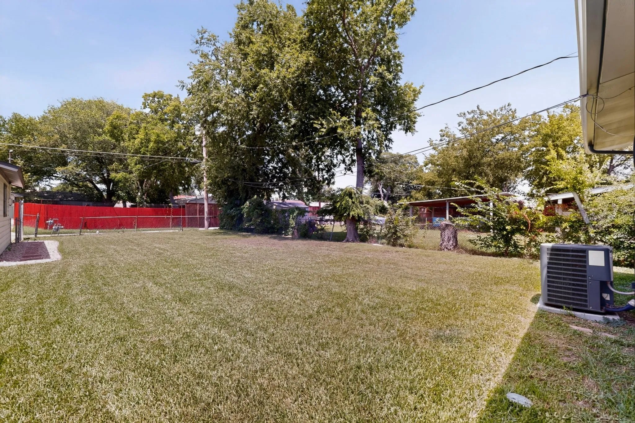 Expansive grassy backyard featuring a red chain-link fence, mature trees, and a visible exterior HVAC unit