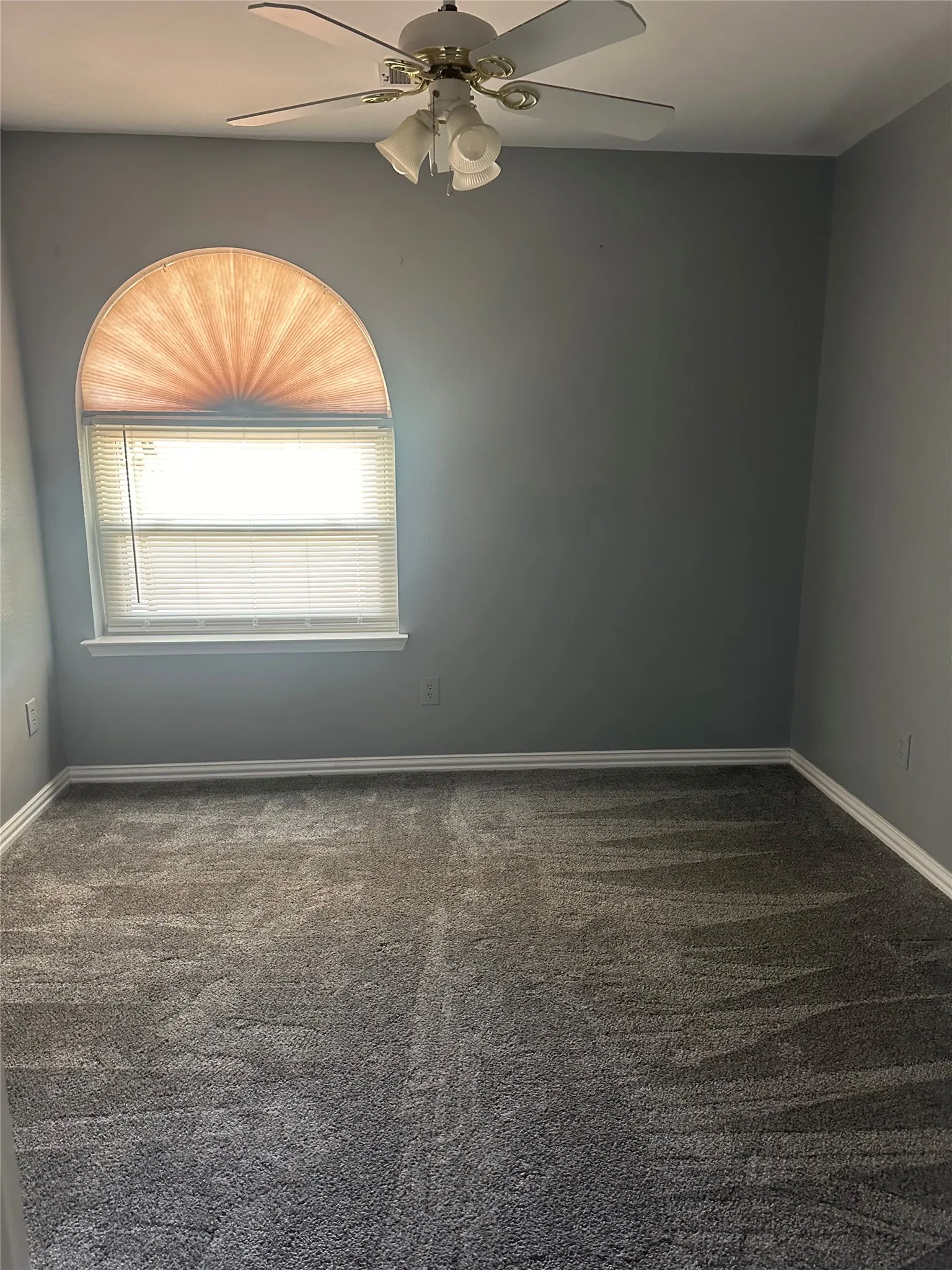 Room featuring a half-moon arched window with pleated blind, ceiling fan with integrated lighting, gray wall paint, and gray carpet flooring with white baseboards