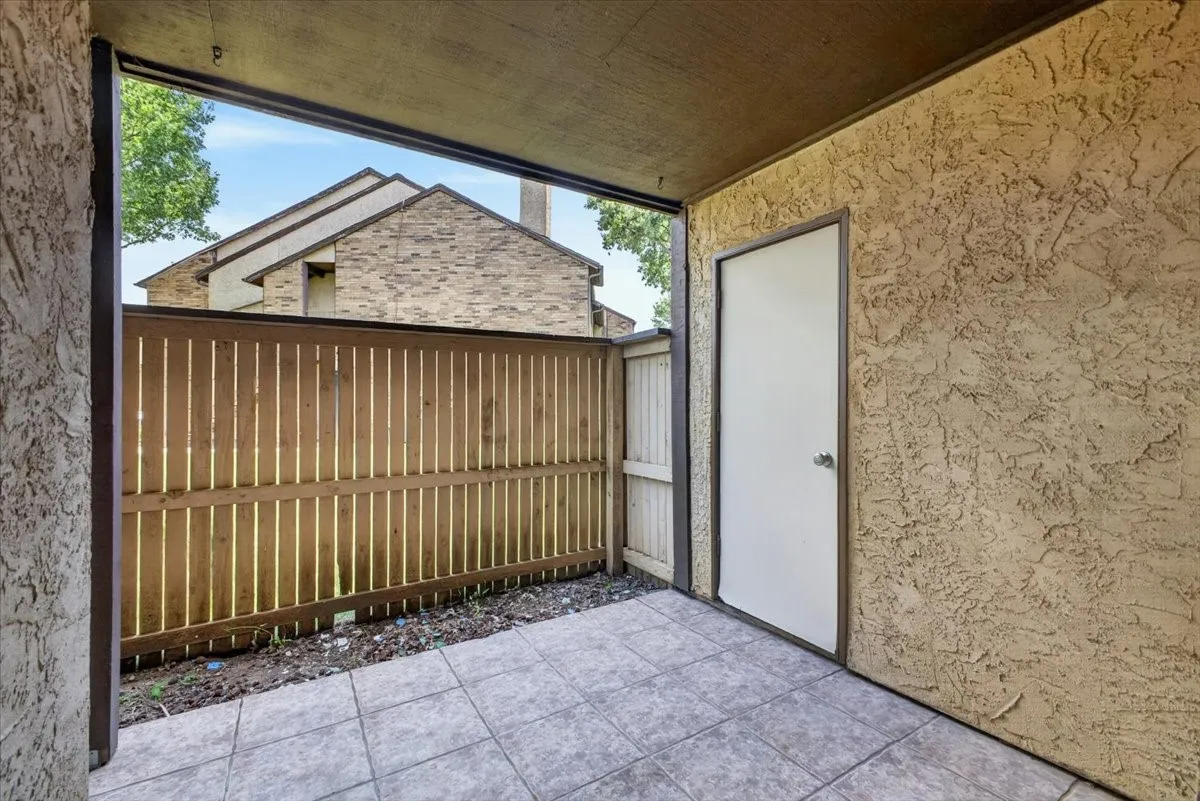 Outdoor covered patio featuring neutral-toned tile flooring, a textured stucco wall, and a wood privacy fence