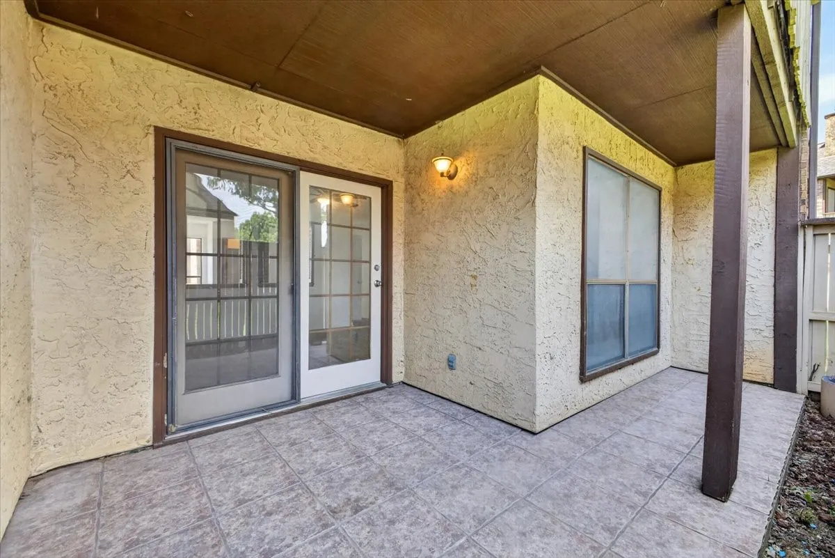 Outdoor covered patio featuring neutral-toned tile flooring, a textured stucco wall, and a wood privacy fence