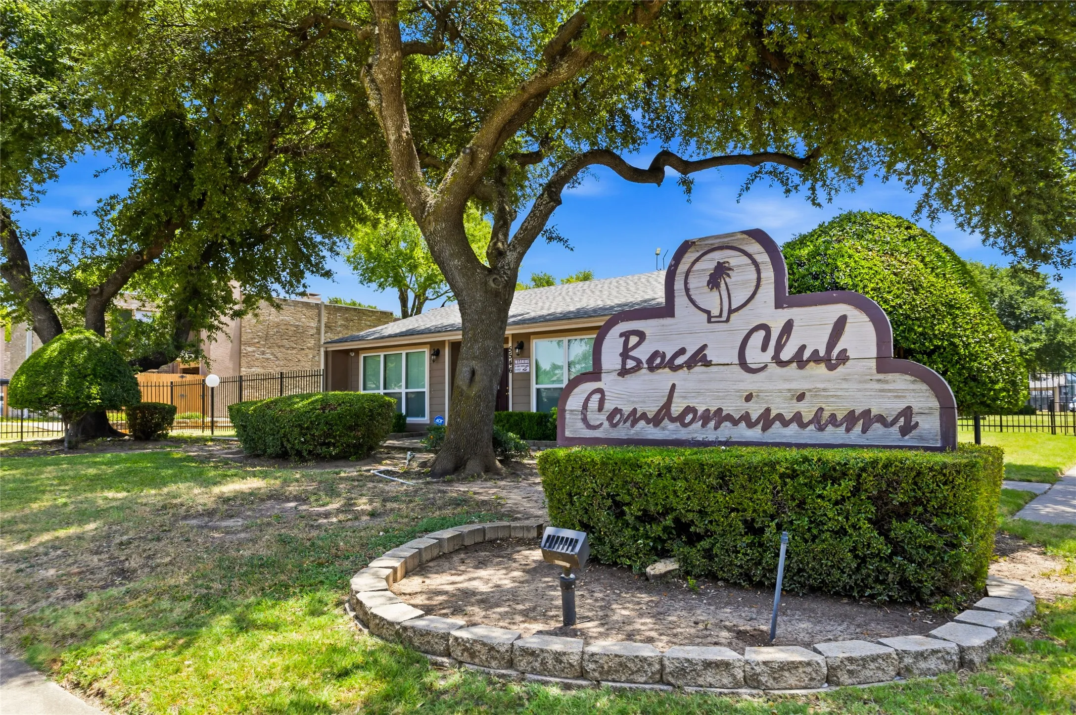 "Boca Club Condominiums" signage with a stone-edged planter, surrounded by mature trees and manicured shrubbery, bordering a grassy area