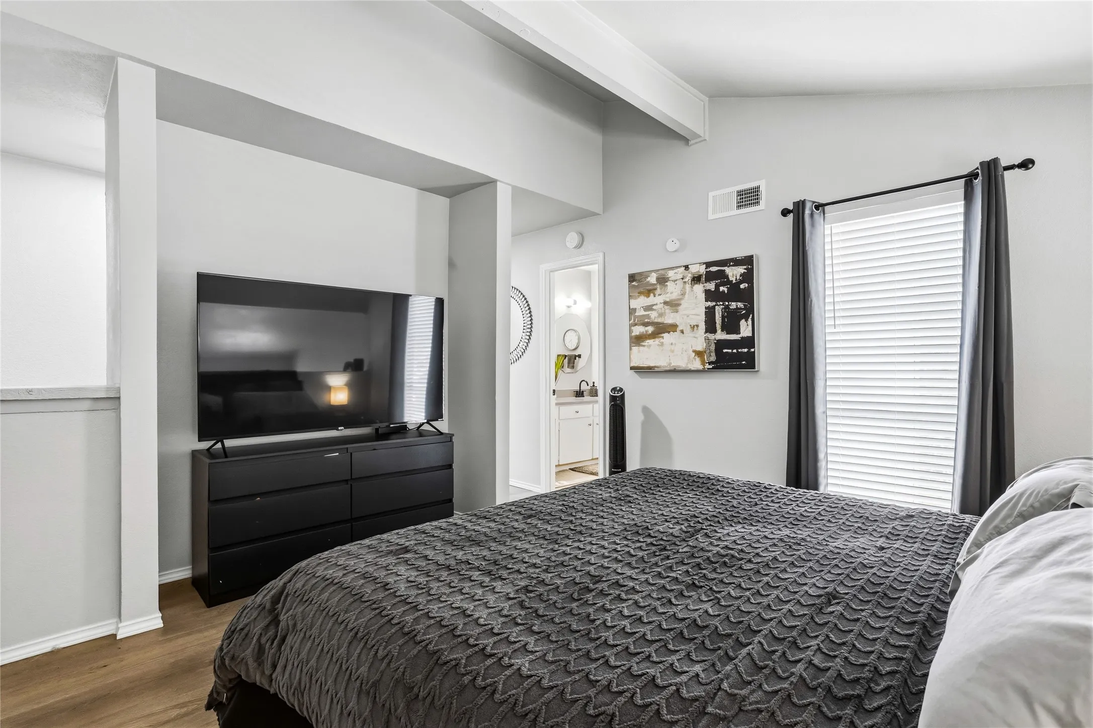 Bedroom featuring wood-finish flooring, a vaulted ceiling with exposed beams, a large window with privacy blinds and dark drapery, and direct access to a bathroom with a white vanity and round mirror