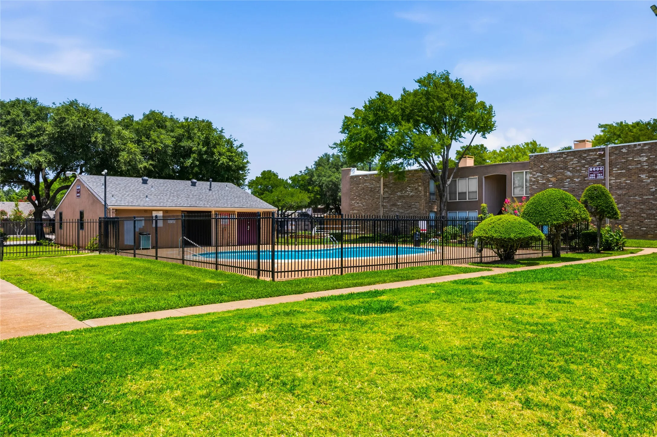 Fenced swimming pool area with a surrounding lawn, mature trees, and a low-rise building featuring brick and stucco finishes