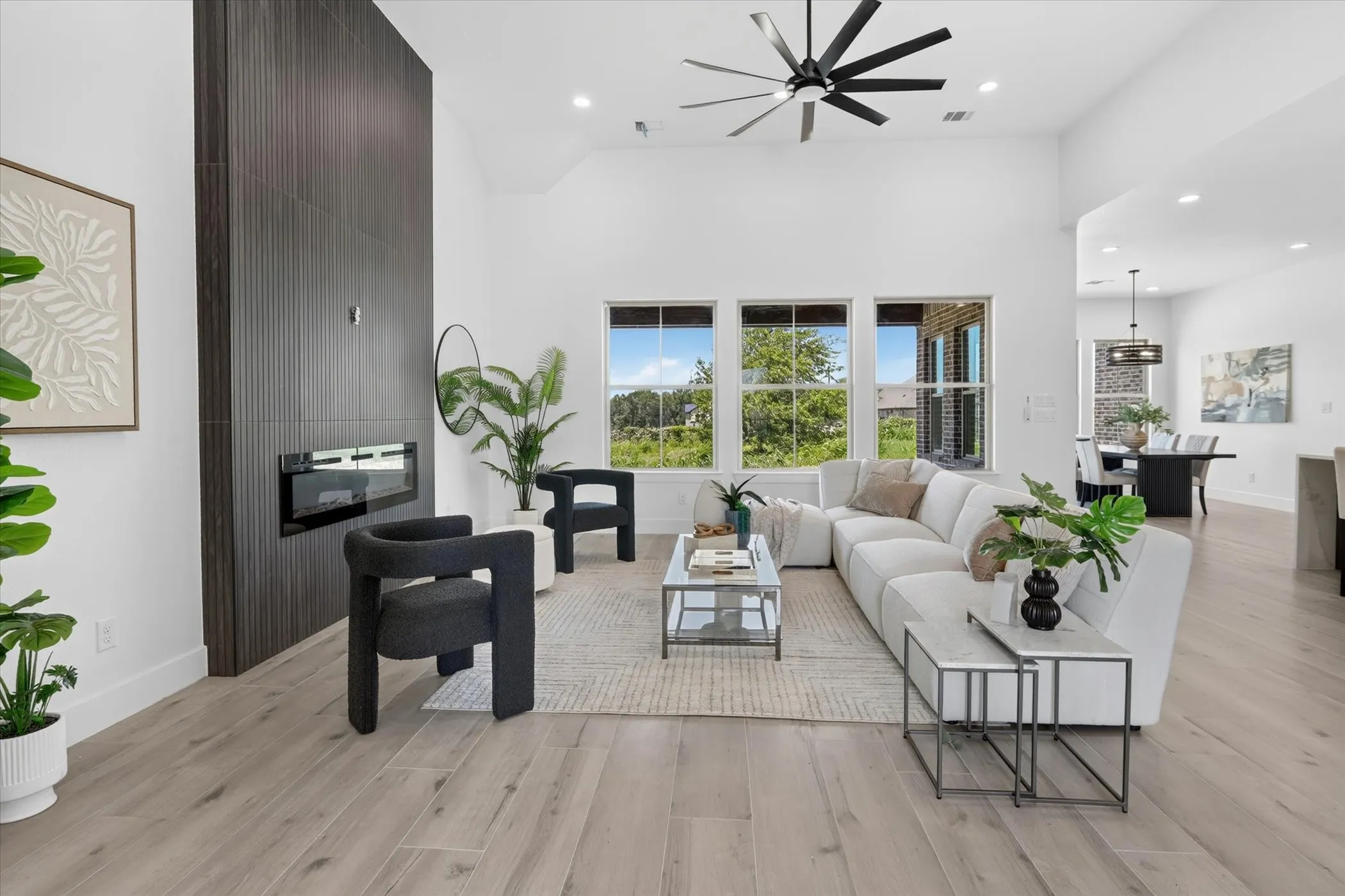 Spacious living area featuring a tall vaulted ceiling, wood-finish flooring, a contemporary fireplace with a dark paneled surround, an oversized ceiling fan, and large windows providing abundant natural light