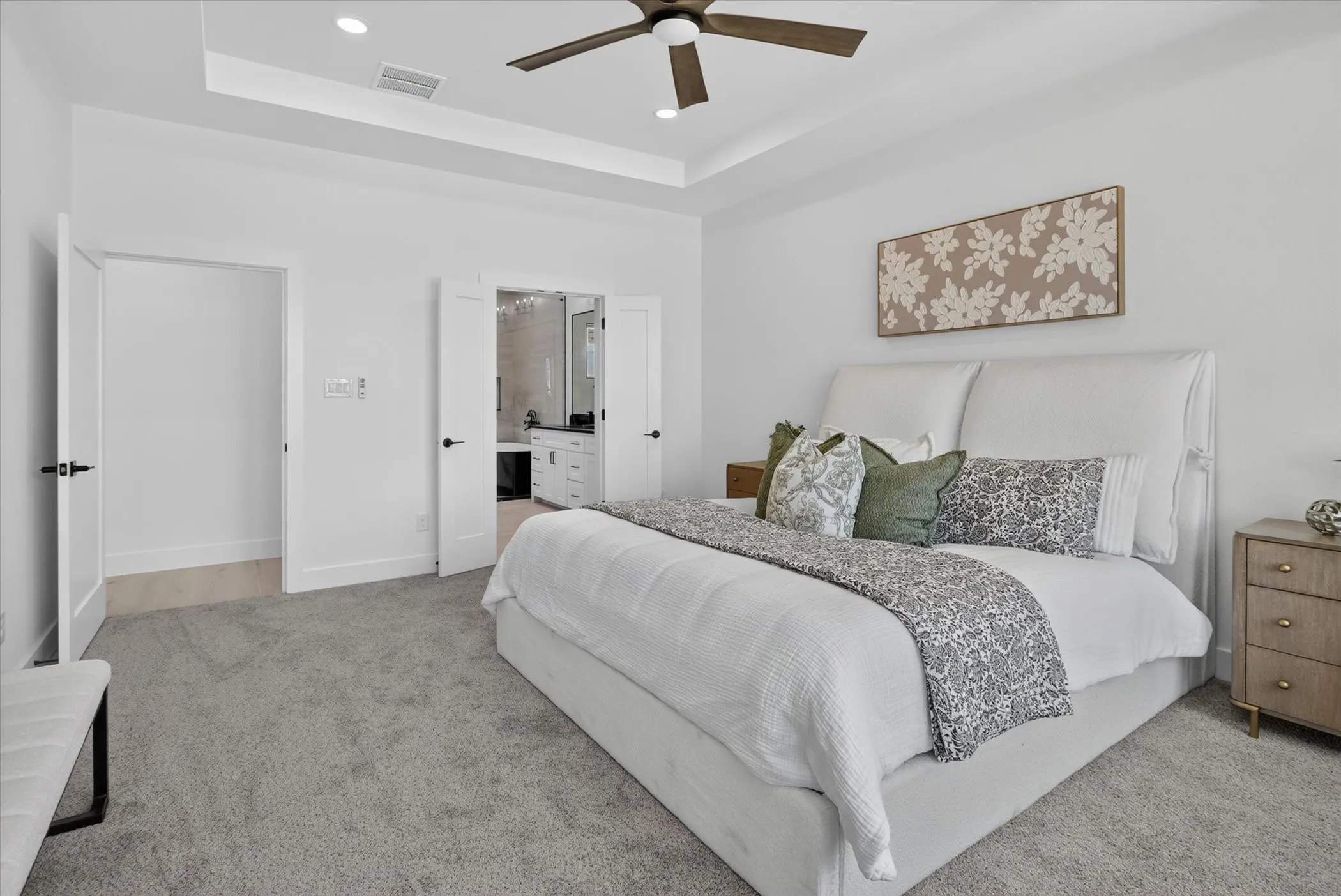 Spacious bedroom featuring light grey carpeting, white walls, a tray ceiling with recessed lighting, and a dark wood-finish ceiling fan