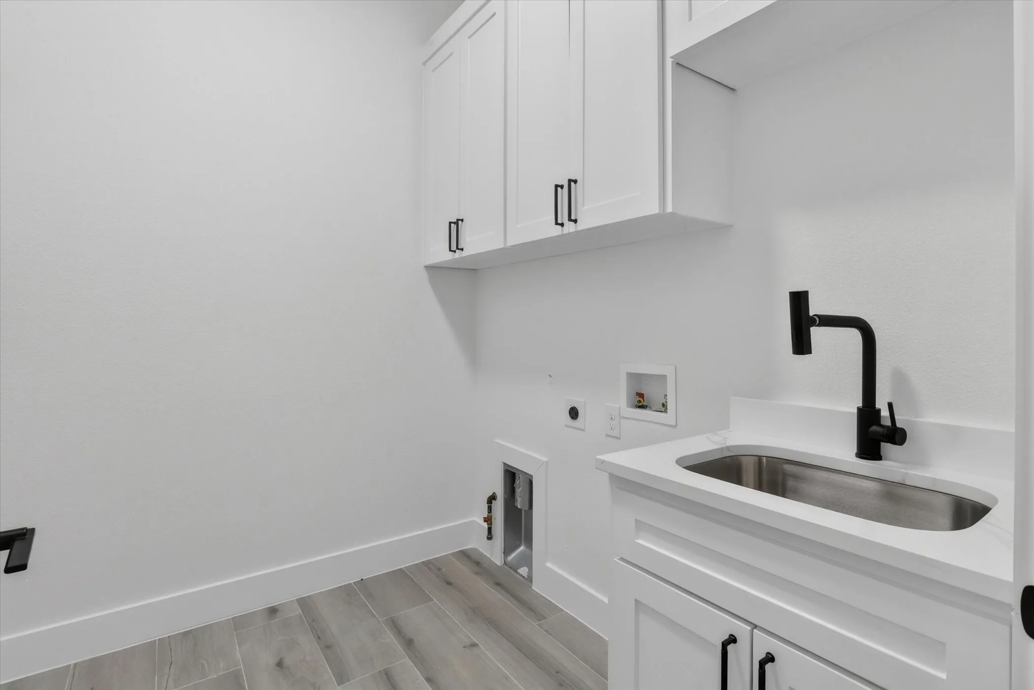 Utility room featuring a built-in sink with a matte black faucet, white cabinetry with black hardware, and wood-finish flooring
