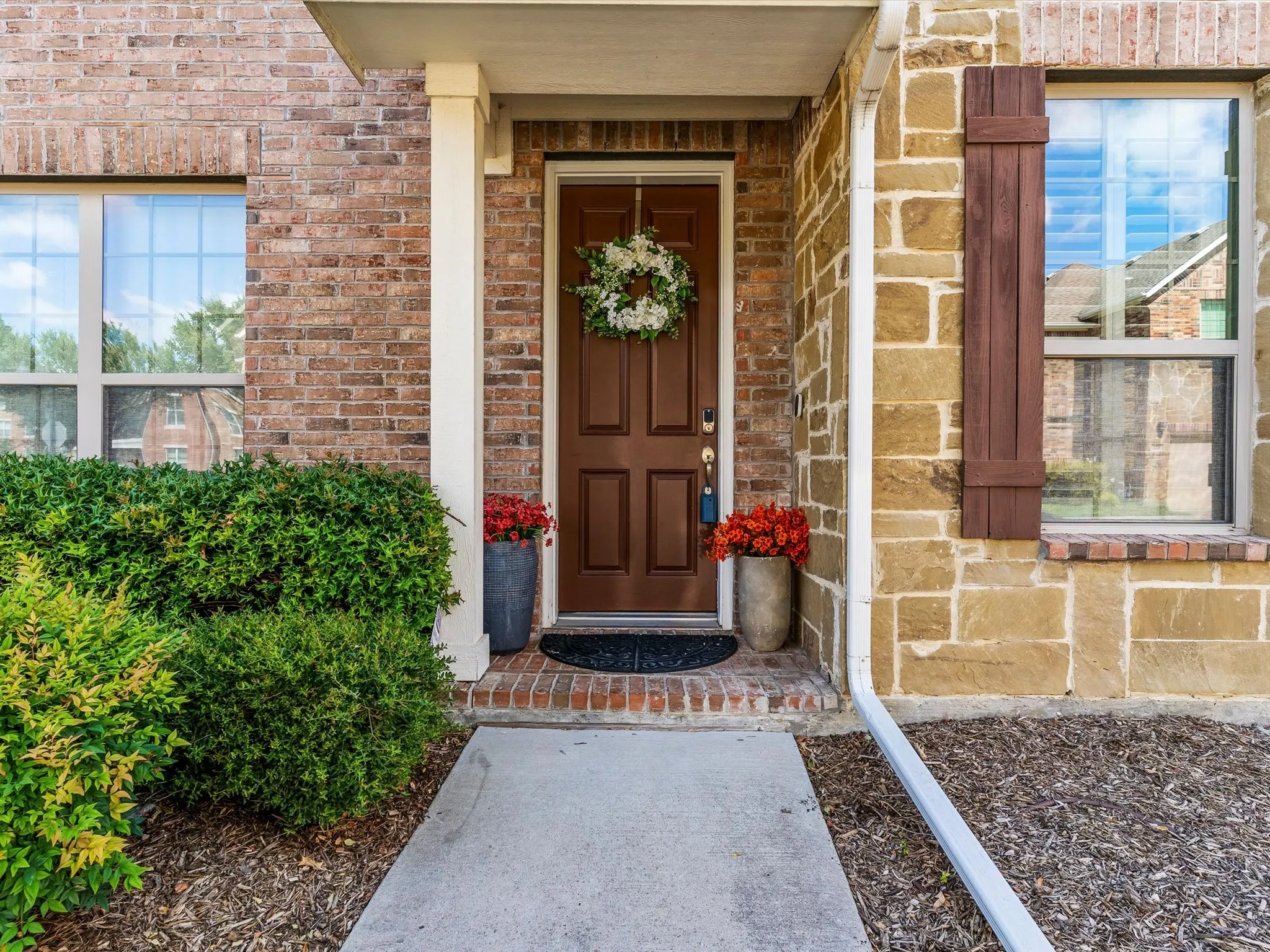 Entryway featuring a paneled front door, mixed material facade with brick and stone, decorative window shutters, and manicured shrubbery