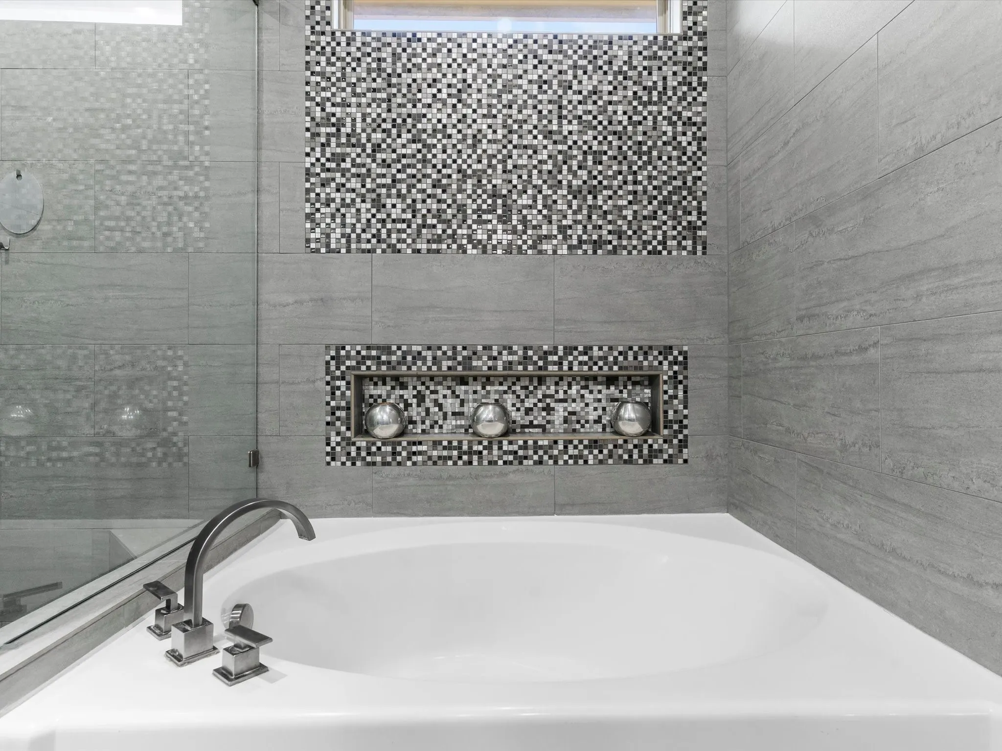 Soaking tub with a brushed nickel Roman faucet, complemented by gray large-format wall tiles and mosaic accents