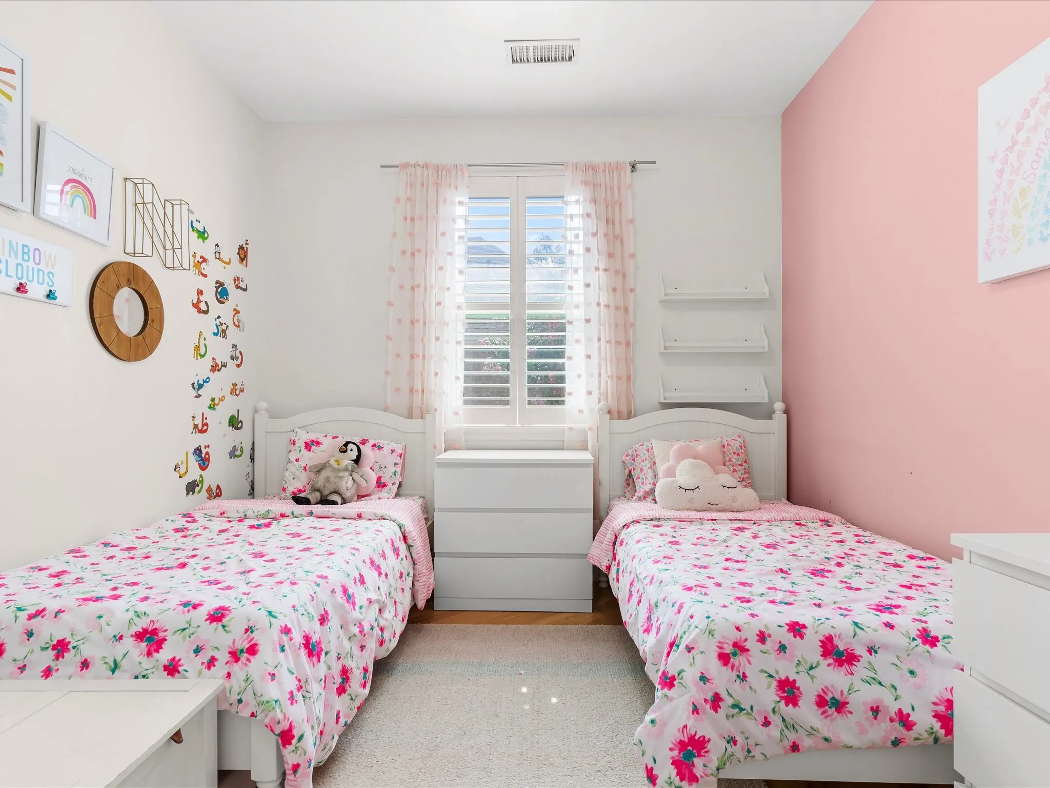 Dual white twin beds with headboards, hardwood-style flooring, a white area rug, and a window with white plantation shutters