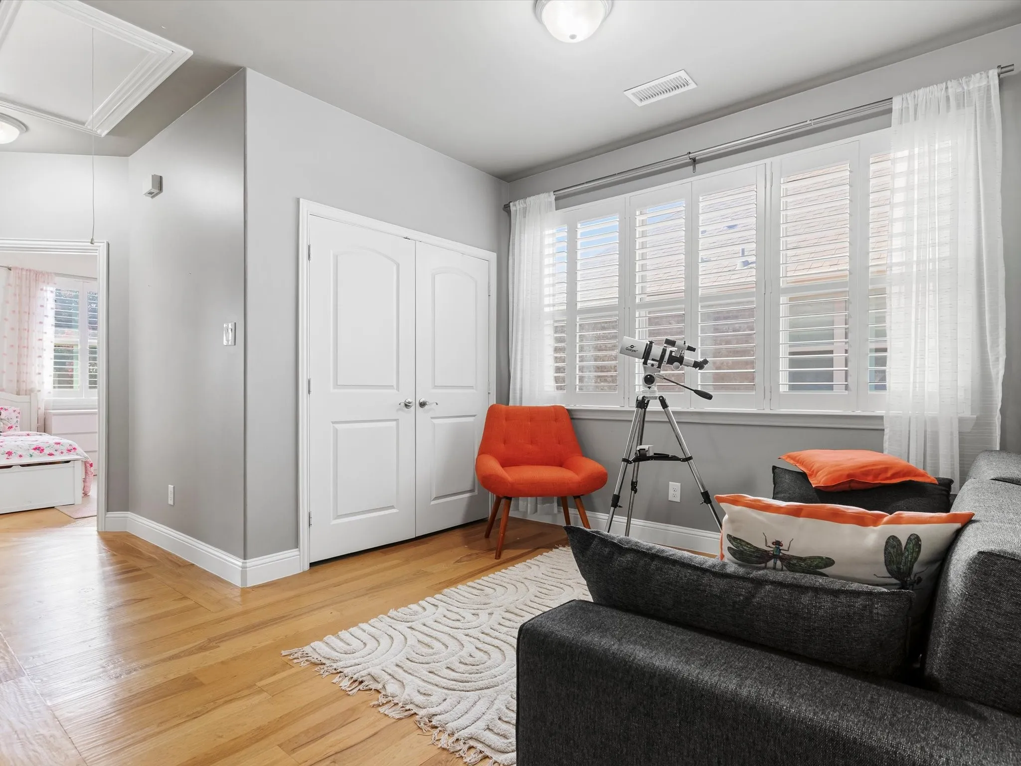 Hardwood flooring throughout featuring white baseboards, a double-door closet with paneling, and a large window with white plantation shutters and sheer curtains