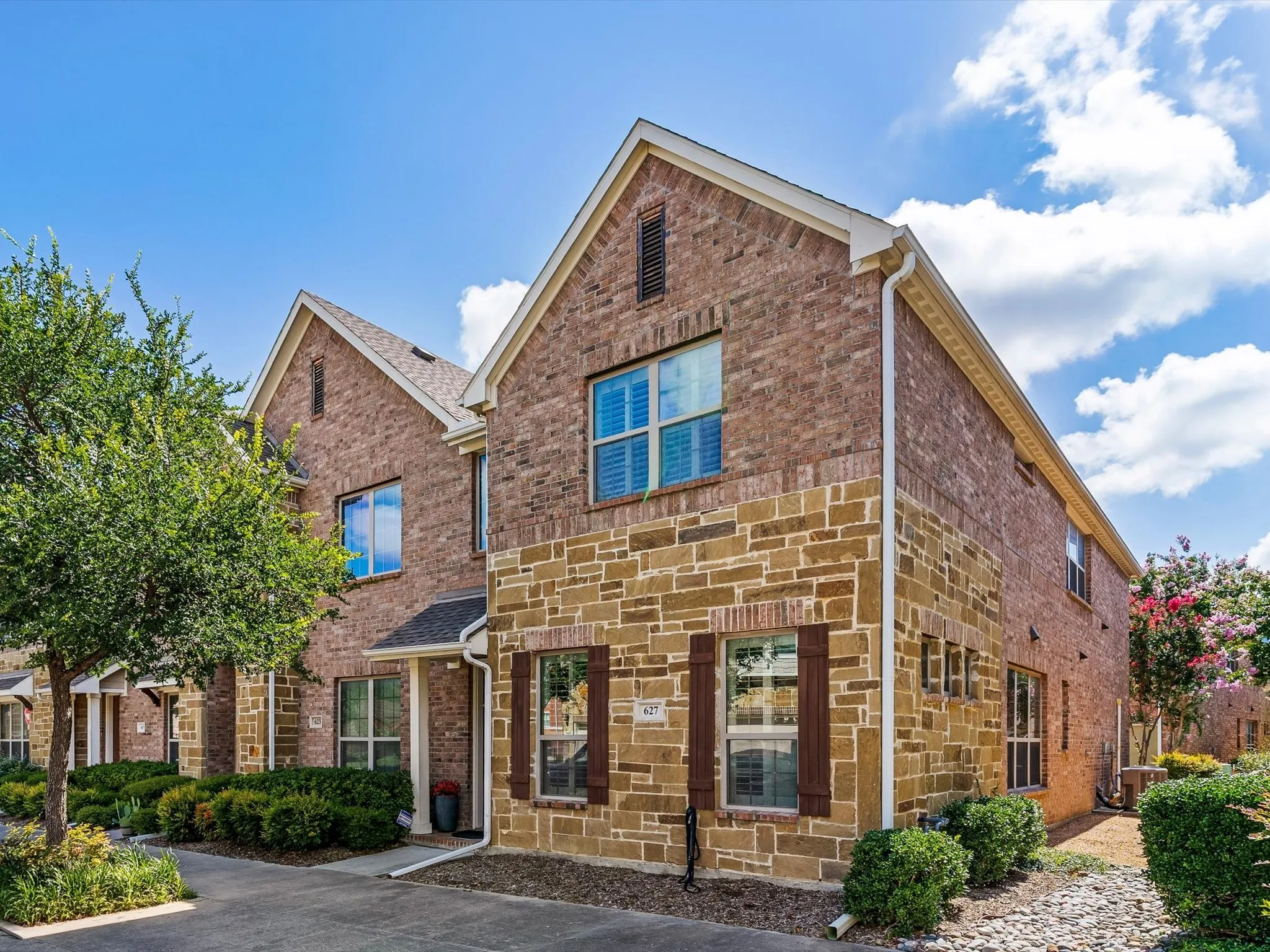 Brick and stone facade townhome featuring gabled rooflines, multiple windows with shutters, and mature landscaping