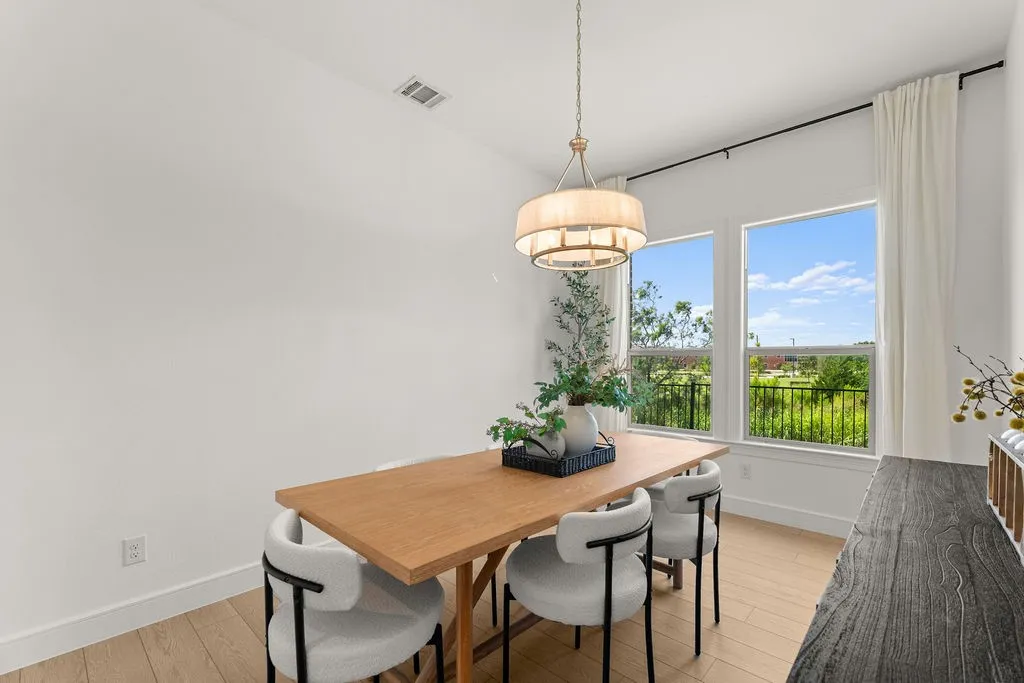 Spacious room with light wood-finish flooring, featuring a tiered drum pendant light fixture, three large windows, white baseboards, and a white wall finish