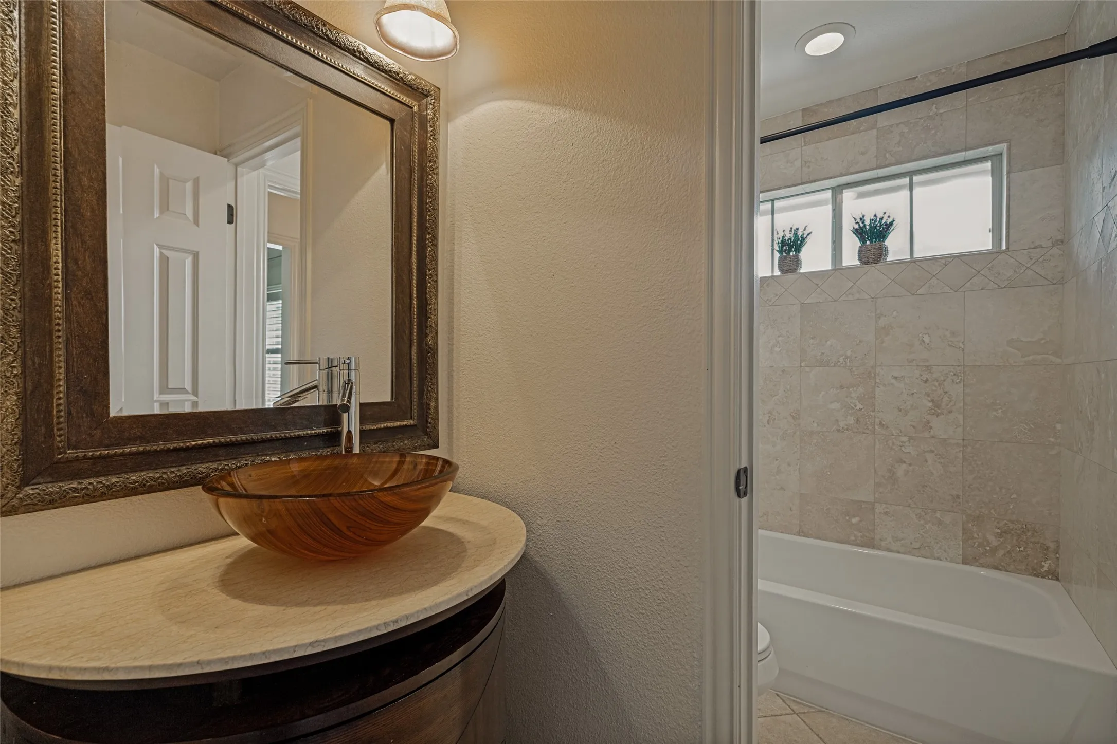 Each side of the Jack and Jill bath has the same decorative features.  Influenced by Art Deco Revival style, the  curved wood furniture piece serves as a vanity.  It is topped by a similarly curved polished Calcutta marble counter with a glass vessel sink. A framed mirror complements the vanity and is topped by distinctive lights.  The floor is ceramic tile with a similarity to the travertine surrounding the tub/shower.  A window at the top of the shower lets natural light into the wet area of the bath.
