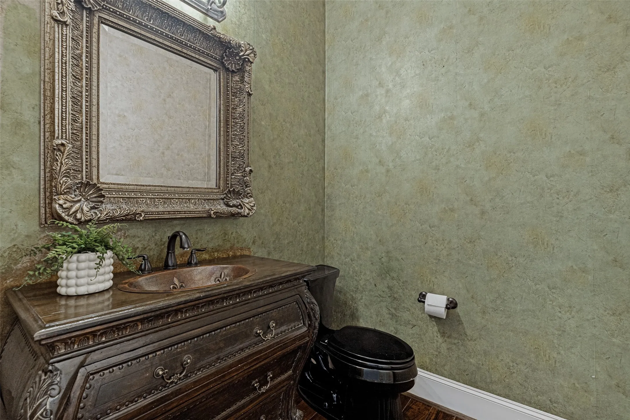 An elegant powder bath is located on the first floor in a side hall off the foyer.  Rich wallpaper with a faux paint look is on the walls.  A handsome bombe chest serves as a vanity with a bronze drop-in sink installed.  An ornate mirror is above.  Wood floors are in the room and decorative lights are above the mirror.