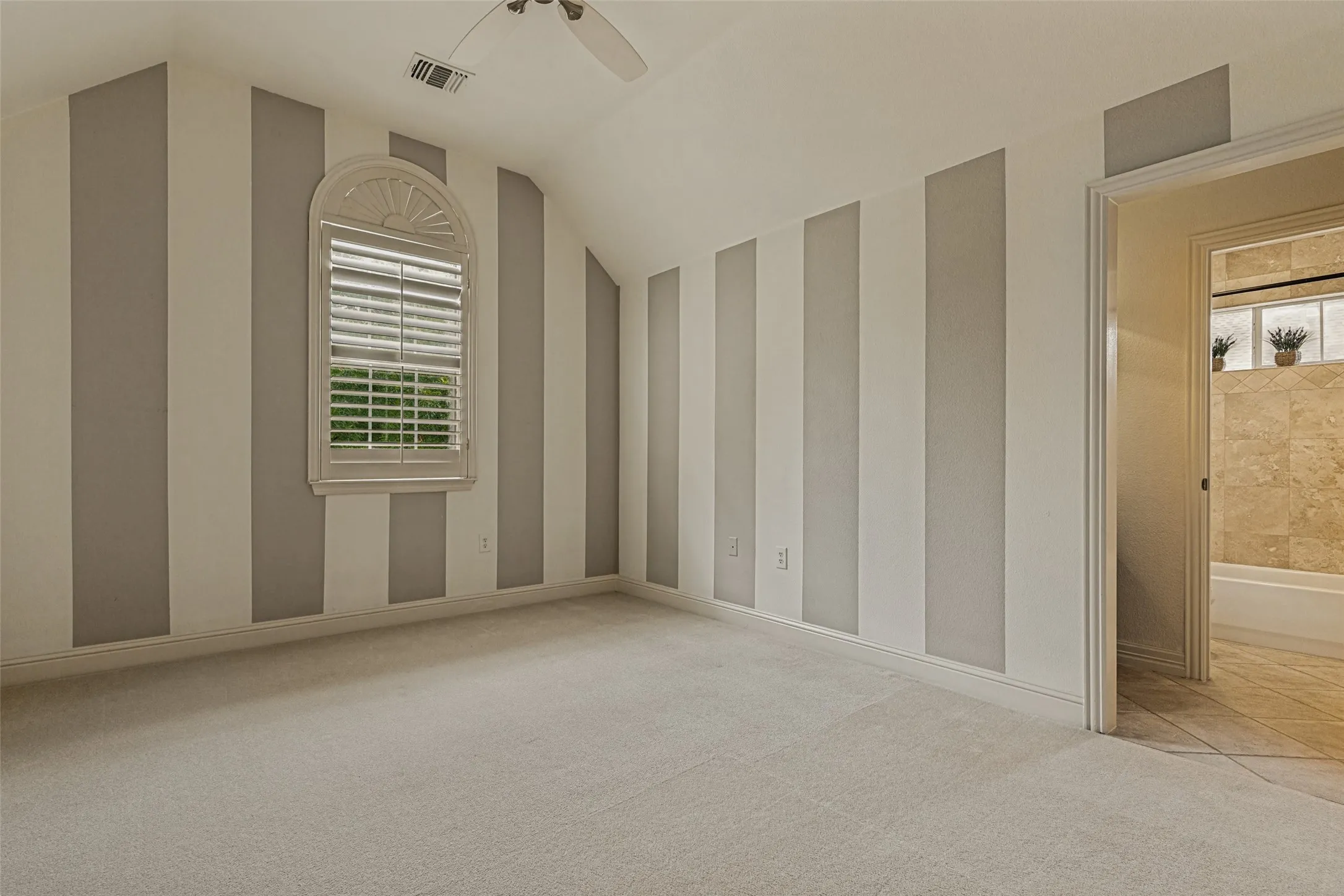 An upstairs bedroom has treetop views of the front of the home.  Jaunty broad light taupe stripes make a clever backdrop for furnishings.  An arched window has shutters, and the raised ceiling sports a breezy fan.  Soft carpet is on the floor.  The room has access to a Jack and Jill bath.