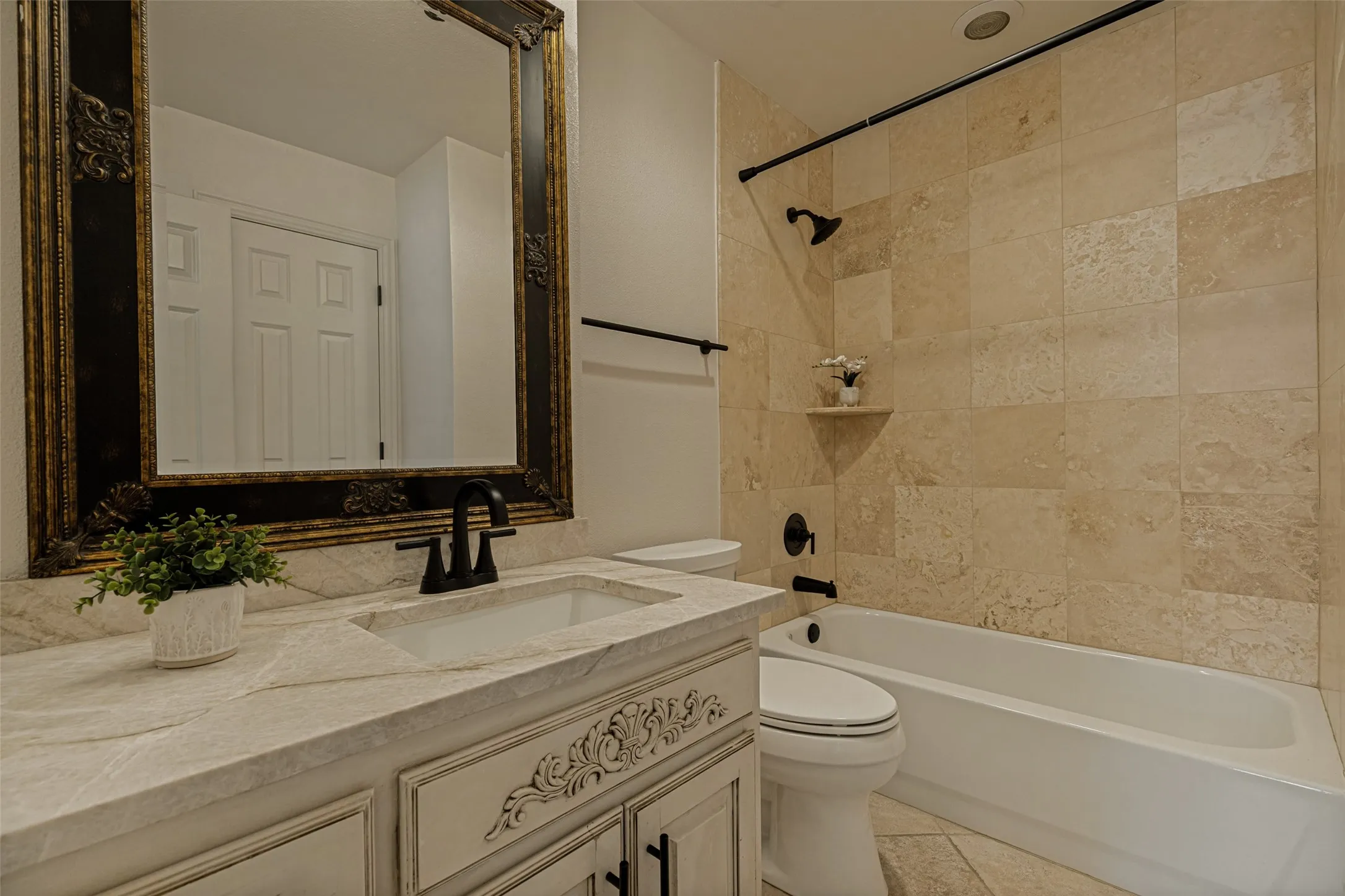 This pretty bathroom was updated with travertine tile on the bath surround.  A glazed finish is on the light colored vanity which is topped by a lovely Calcutta marble countertop.  Touches of French influence are found on the mirror and the carved relief effect on the vanity.  Iron hardware and fixtures are in the bath, and tile with a travertine look is on the floor.  This bath serves as a hall bath and also has direct access to the media/bedroom.