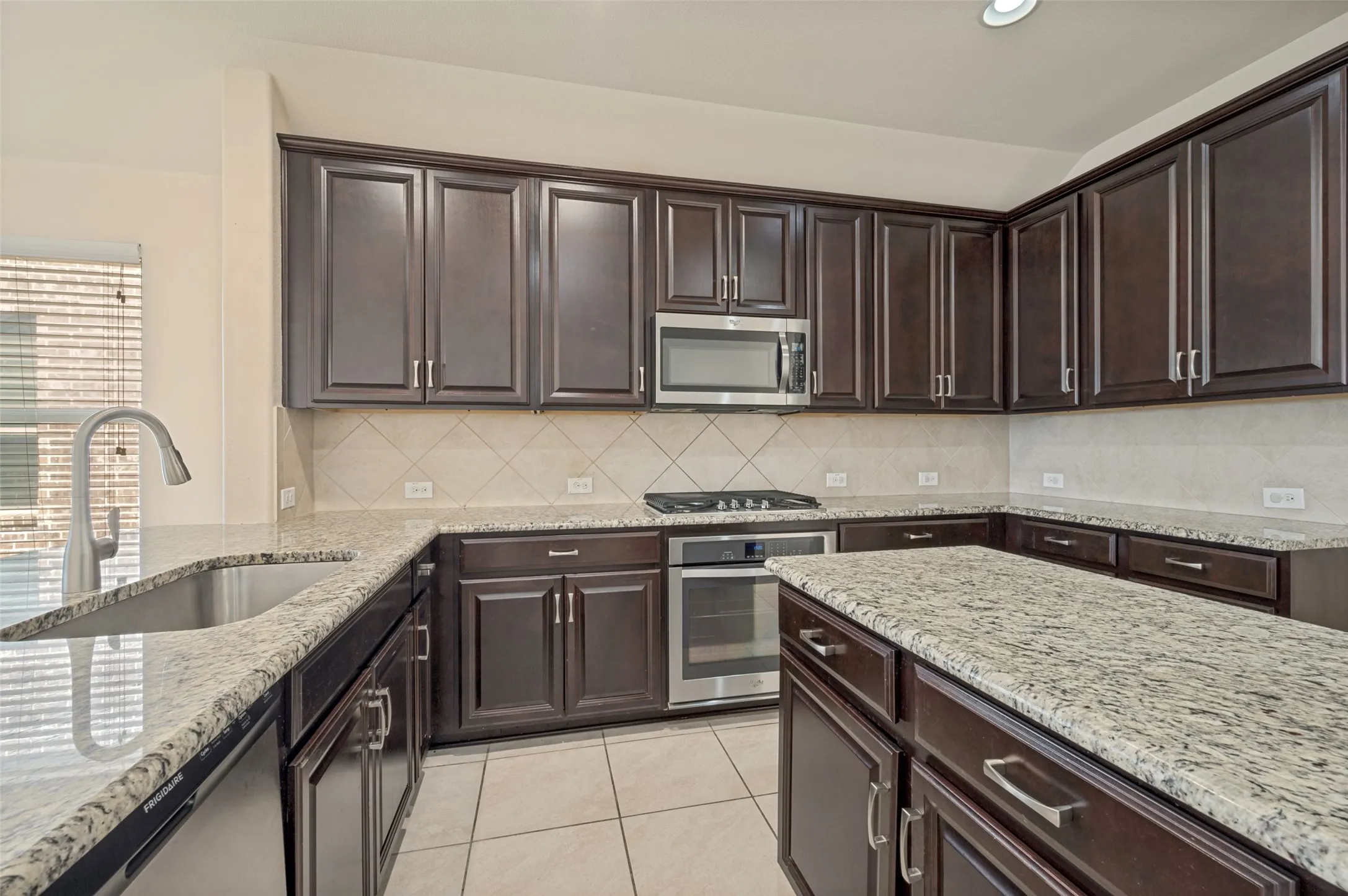 Kitchen featuring extensive dark wood cabinetry, granite countertops, a stainless steel sink with a high-arc faucet, and stainless steel appliances including a built-in microwave and oven