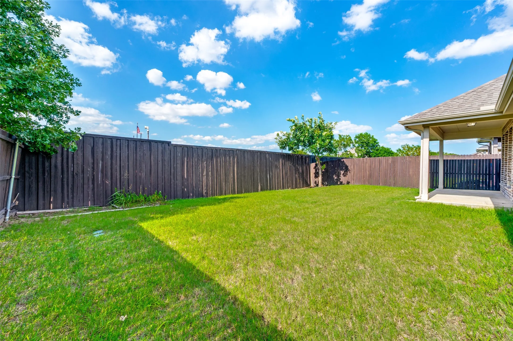 Expansive grassy backyard featuring a tall privacy fence, mature perimeter trees, and a covered patio with columns and a tiled roof