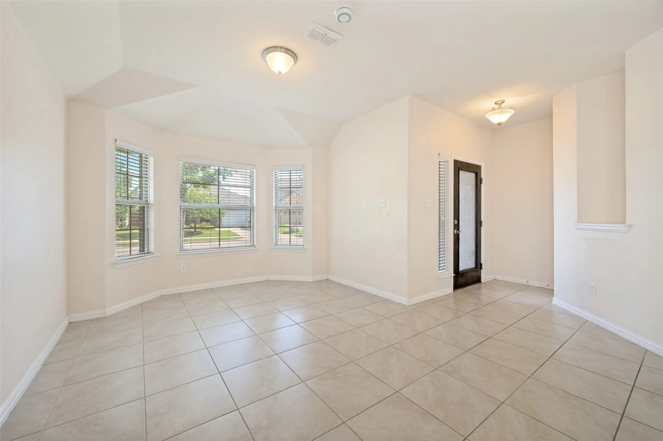 Spacious interior featuring neutral tone walls, large format ceramic tile flooring, a bay window with blinds, and an entryway with a dark-framed door and frosted glass insert