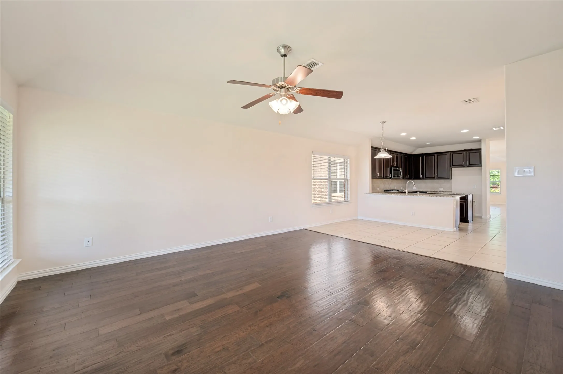 Spacious interior featuring wide-plank wood-finish flooring, a ceiling fan with integrated lighting, and a kitchen with dark cabinetry, an island with pendant lighting, and tile flooring
