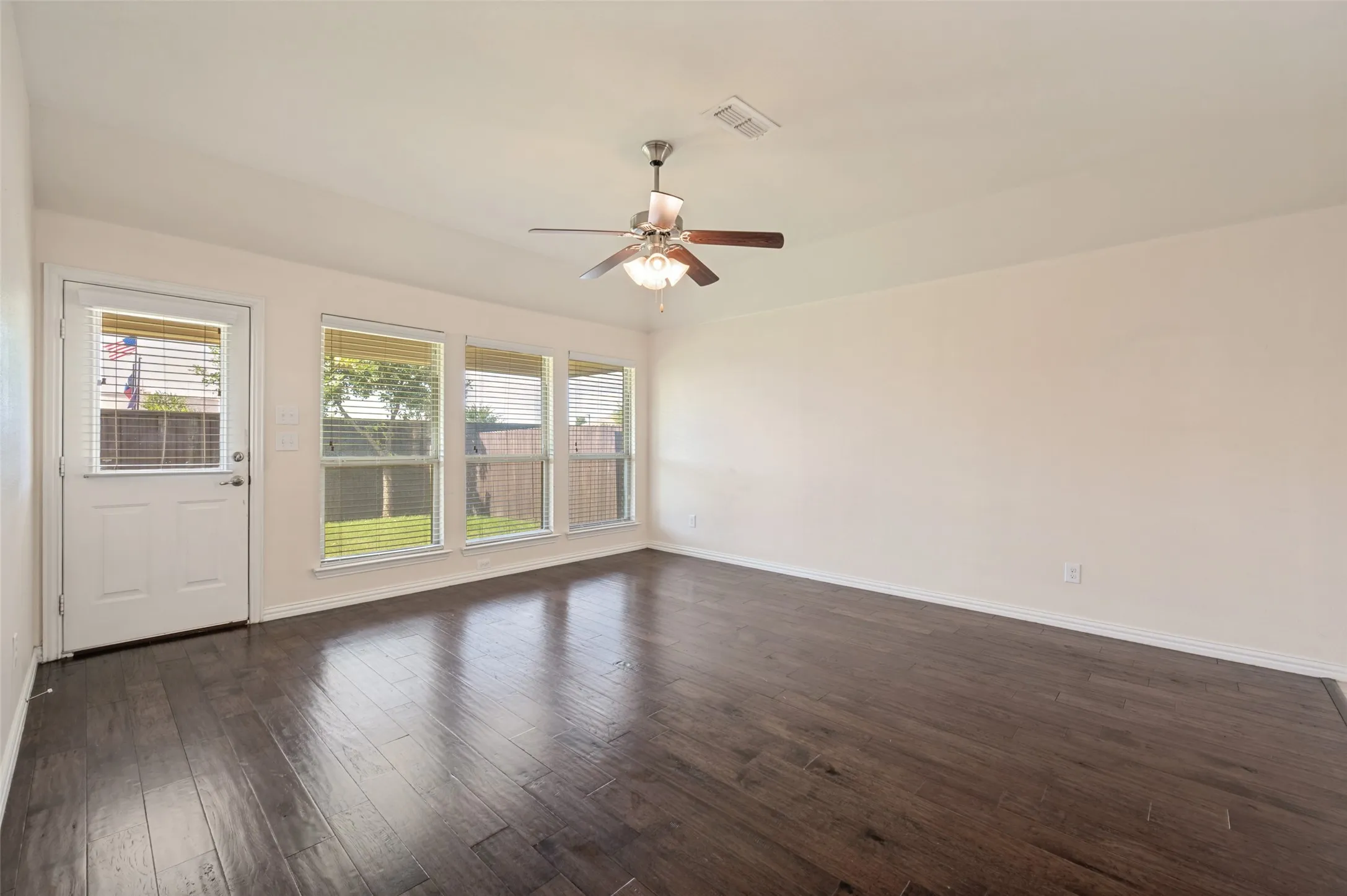 Spacious room featuring dark wood-finish flooring, a wall of multiple windows with blinds, a white door with a window, and a ceiling fan with integrated lighting