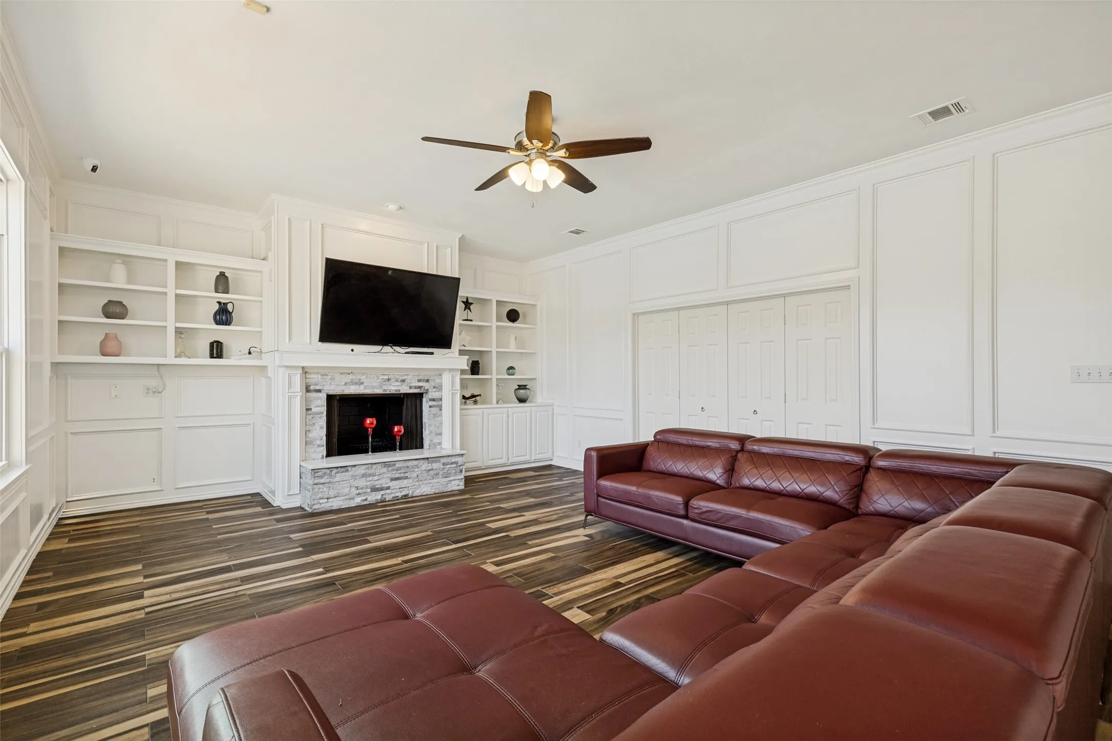 Living area featuring a stone-clad fireplace with mantel, built-in shelving, wood-finish flooring, a ceiling fan, and extensive white wainscoting