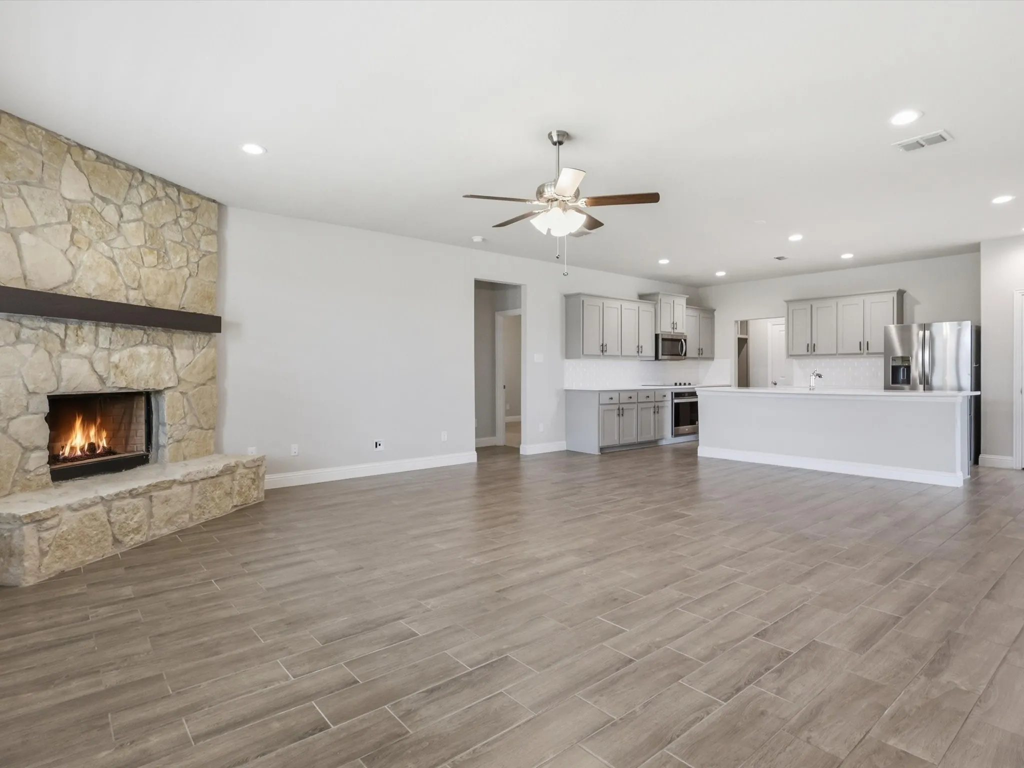 Open-concept living area featuring wood-finish flooring, a stone-surround fireplace with a dark wood mantel, recessed lighting, and a ceiling fan