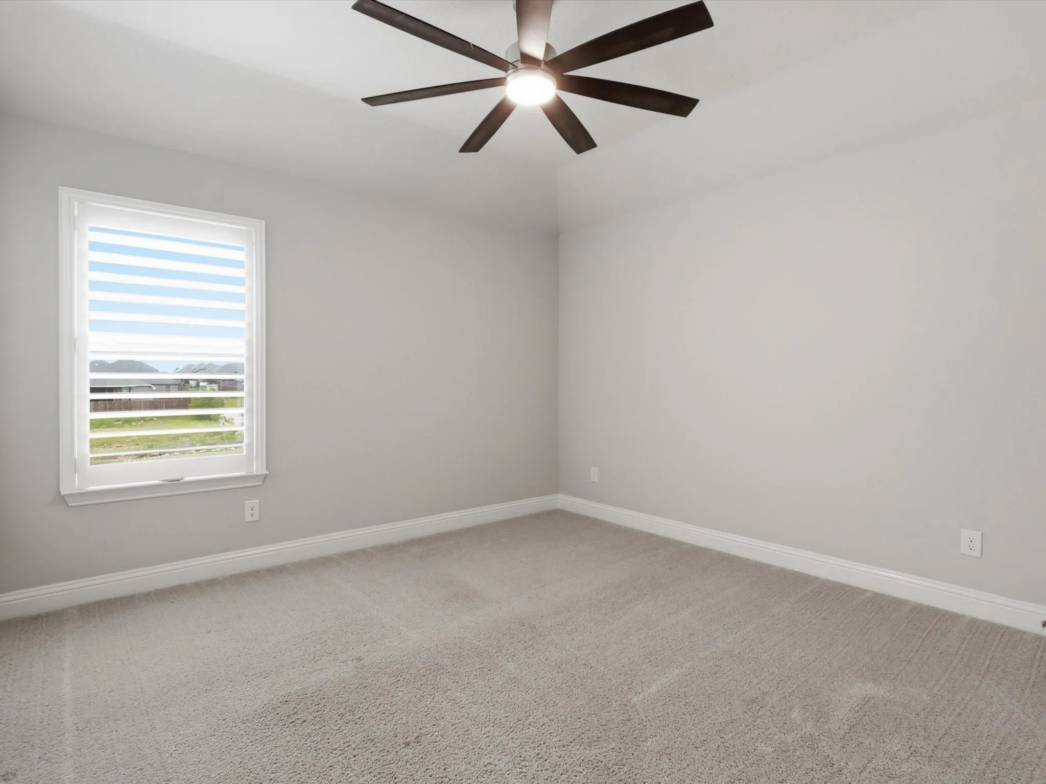 Spacious room featuring neutral carpeting, light gray walls, a white-framed window with blinds, and a dark-toned ceiling fan with integrated lighting
