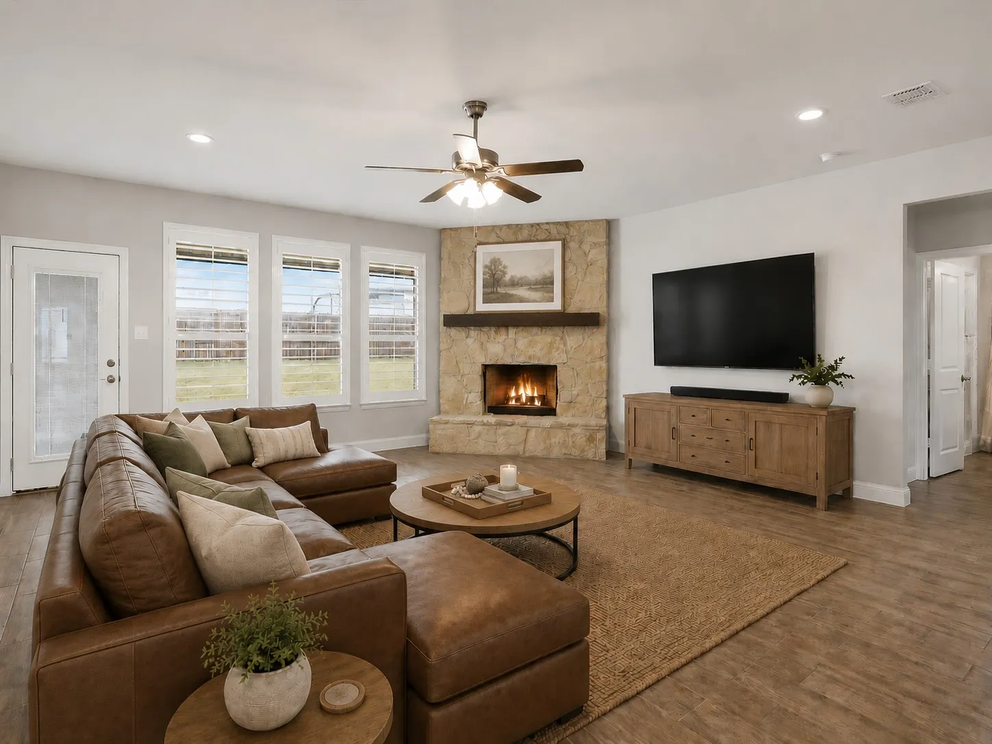 Spacious living area featuring a corner stone fireplace with a wood mantle, wood-finish flooring, a ceiling fan, and multiple windows