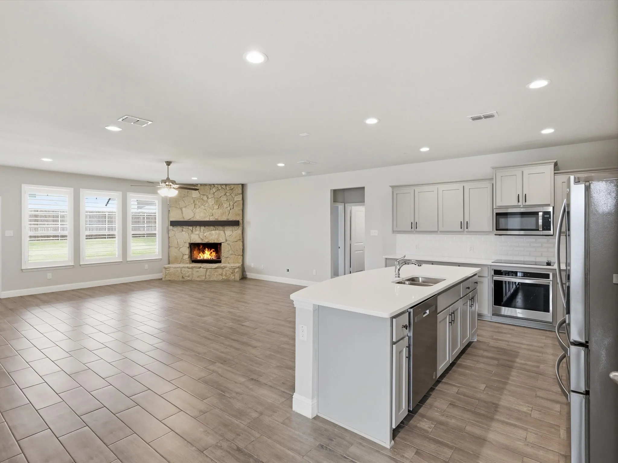 Open-concept living space featuring wood-finish flooring, a stone-faced fireplace with a wood mantle, and a kitchen island with an undermount sink and stainless steel appliances