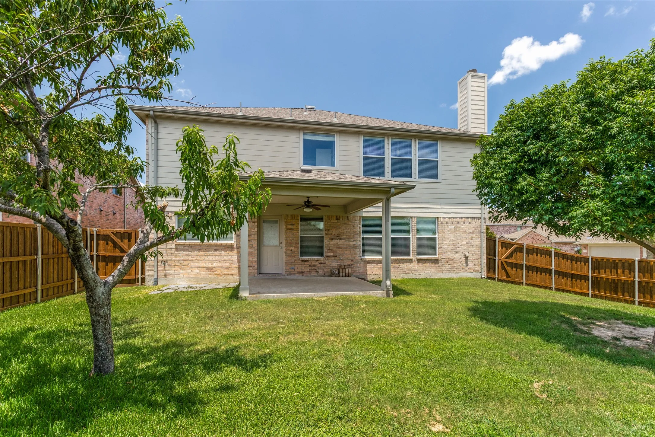 Expansive fenced backyard featuring a covered patio with a ceiling fan, a lush lawn, mature trees, and a brick and siding exterior