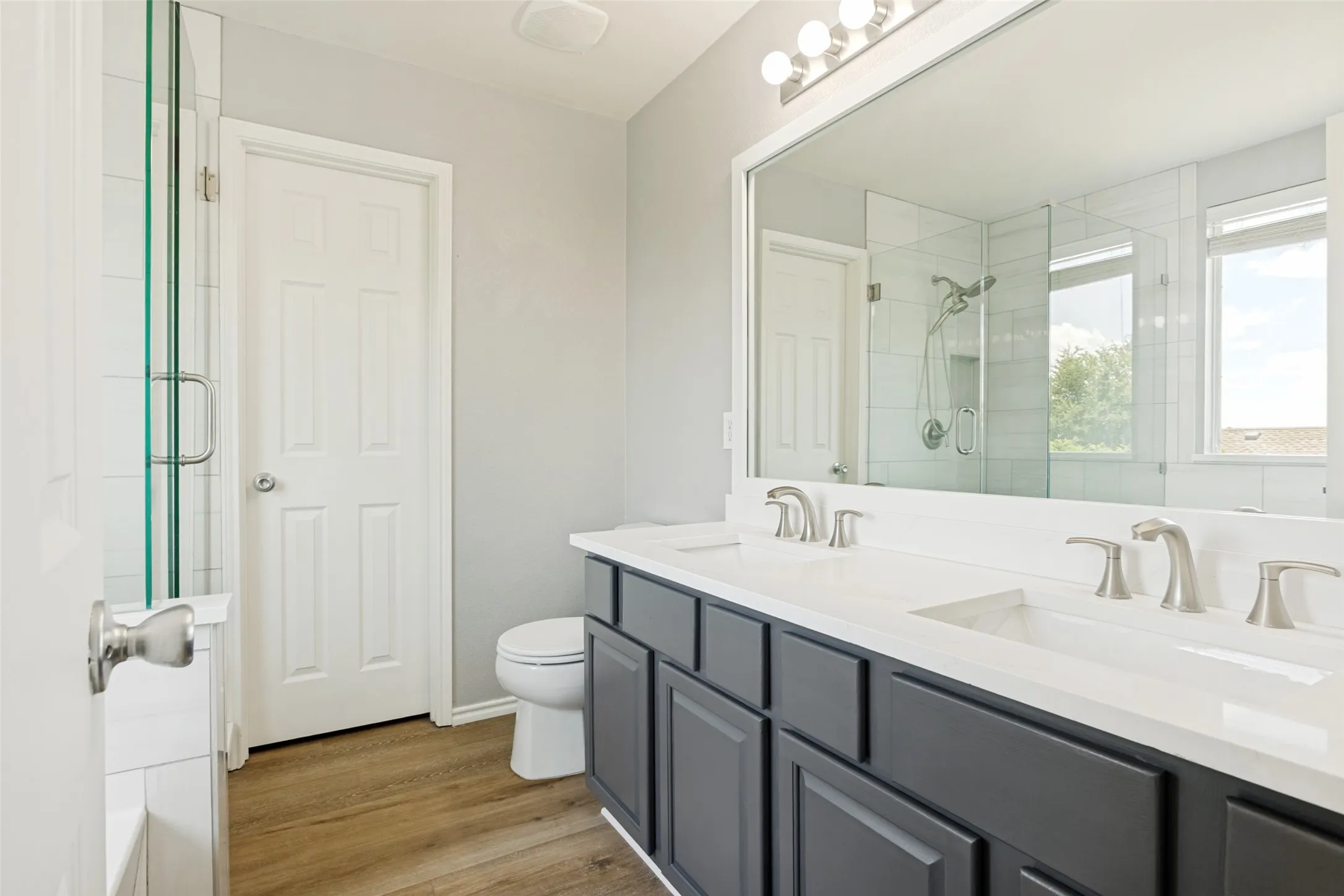 Bathroom featuring a dual vanity with dark cabinetry and white countertops, a large framed mirror, and wood-finish flooring