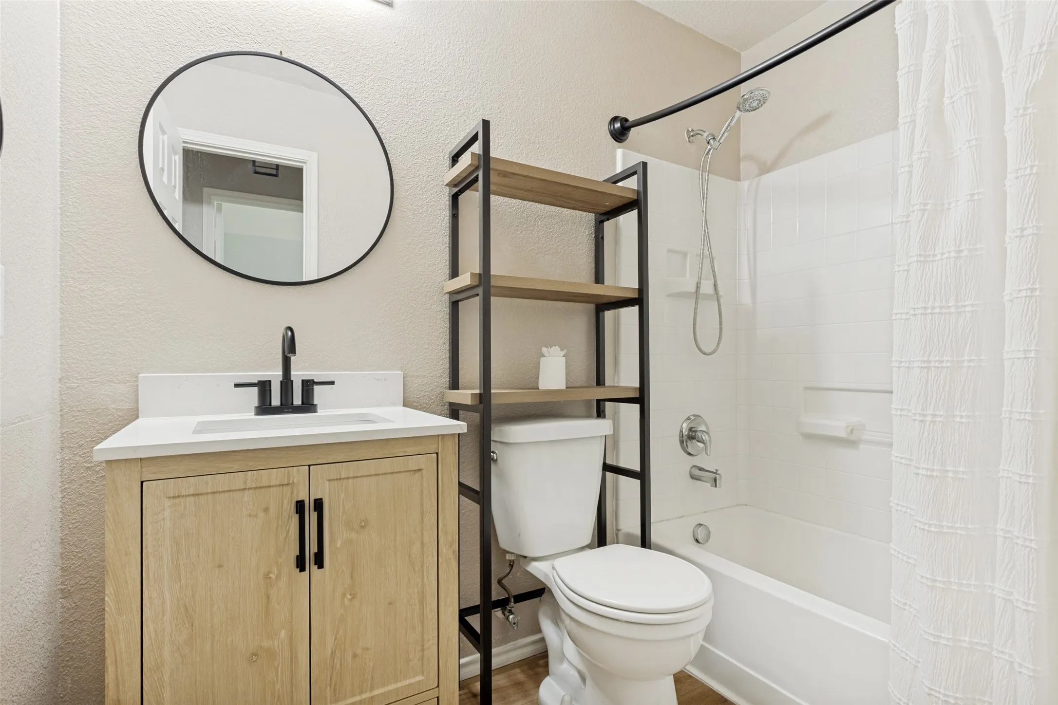 Bathroom featuring a light wood-finish vanity with a white countertop and matte black faucet, a round black-framed mirror, a white toilet with an industrial-style shelving unit, and a white bathtub with a shower head and curved shower rod
