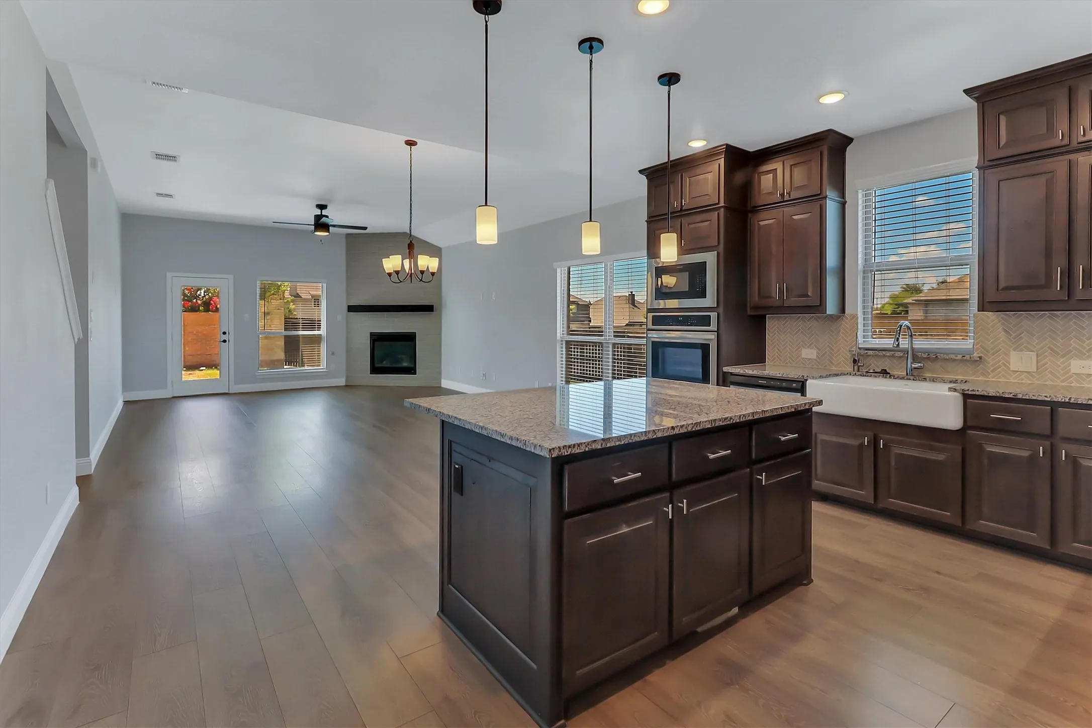Contemporary kitchen featuring an island with a granite countertop, a farmhouse sink, stainless steel built-in appliances, dark wood cabinetry, and a patterned tile backsplash