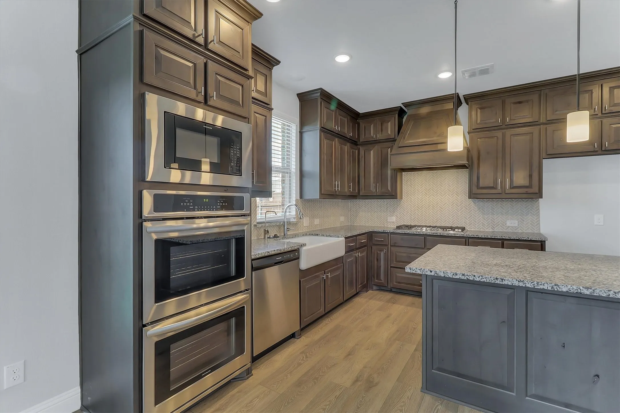 Gourmet kitchen featuring a built-in stainless steel oven and microwave stack, wood-finish flooring, a farmhouse sink, granite countertops, and a full-height tile backsplash
