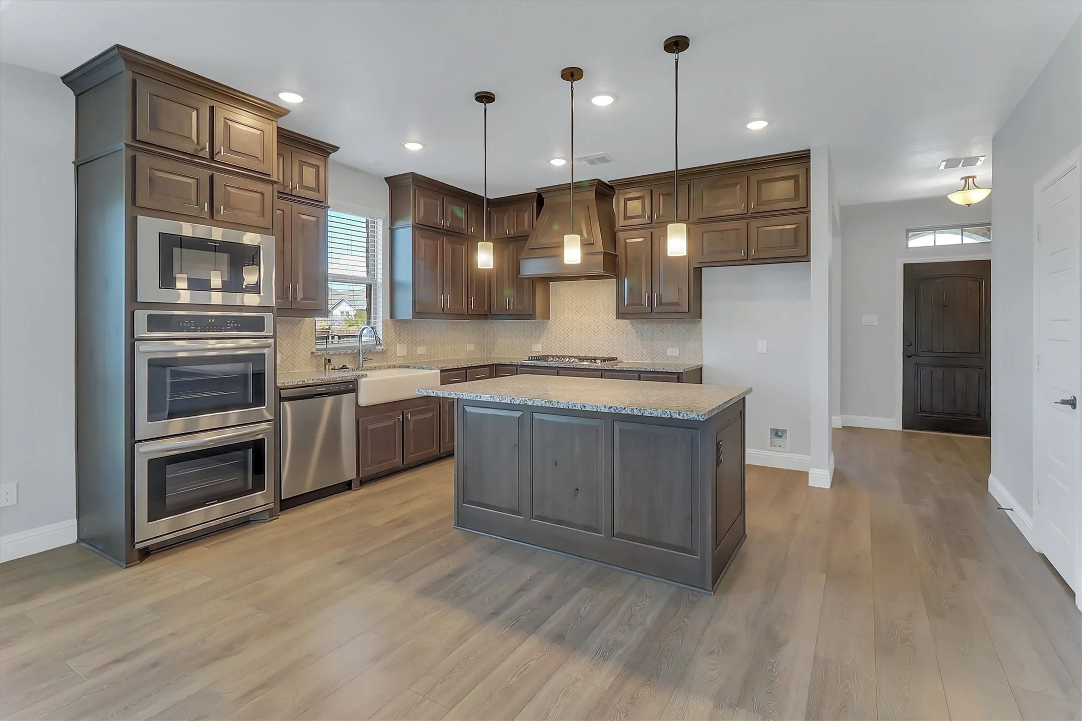 Kitchen featuring wood-finish flooring, dark wood cabinetry, stainless steel appliances including double ovens, a white apron sink, and a center island with pendant lighting