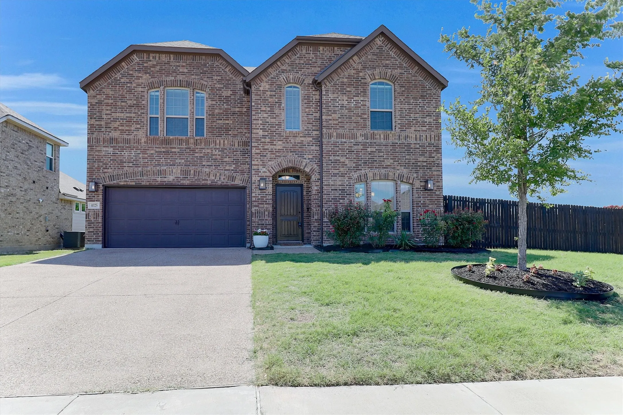 Two-story brick exterior featuring a two-car garage, arched entryways, multiple windows, and a landscaped front yard with a tree